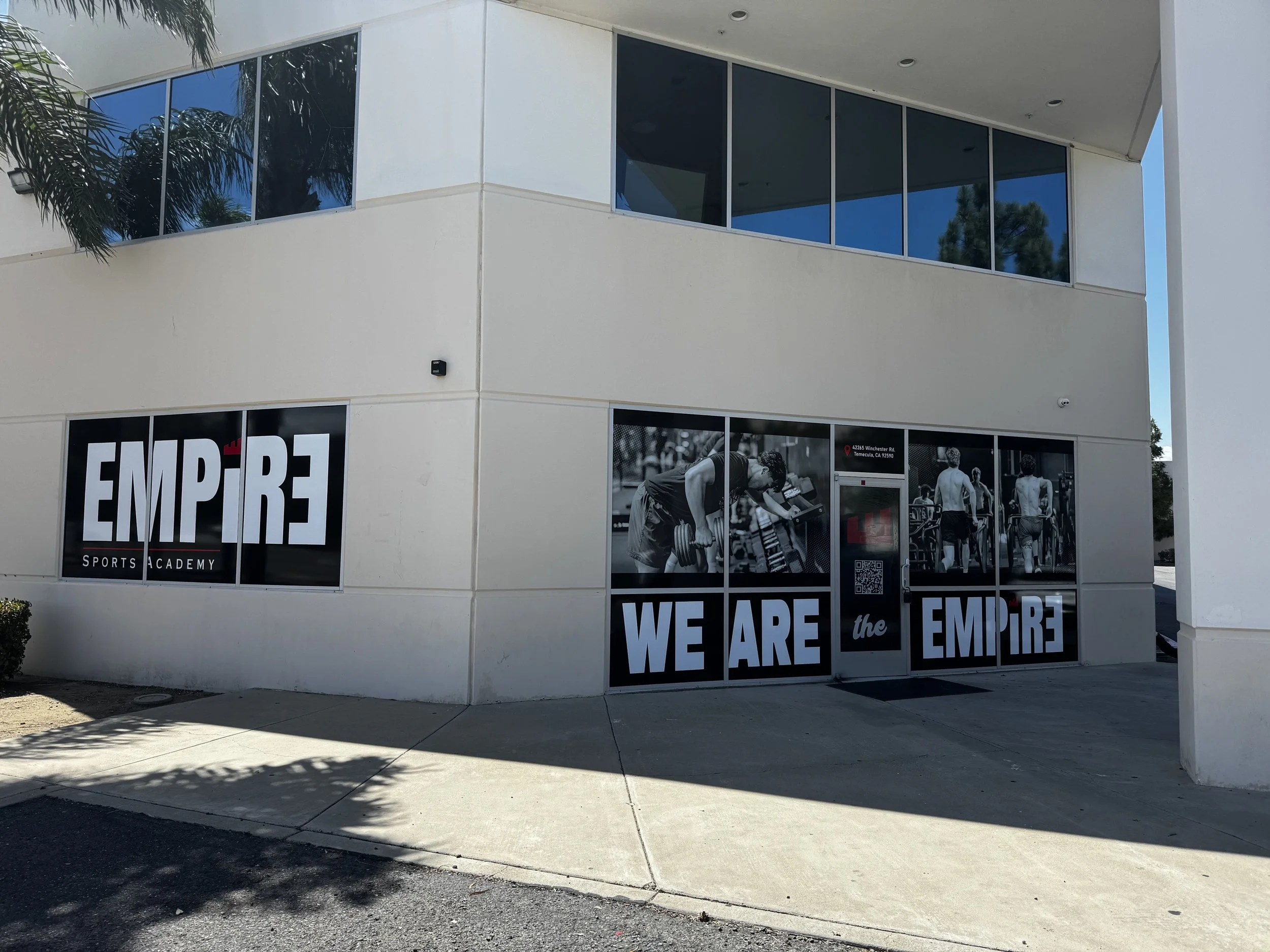 Exterior of a white building with large windows, advertising 'EMPIRE Sports Academy' with photos of people working out, and the slogan 'WE ARE the EMPIRE' on the windows.
