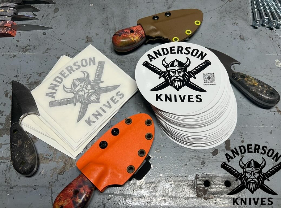 Stickers and knives on a grey work surface, including a stack of stickers with a Viking warrior logo and the text "Anderson Knives," a partially visible orange knife sheath, and two knives with black and wood handles.
