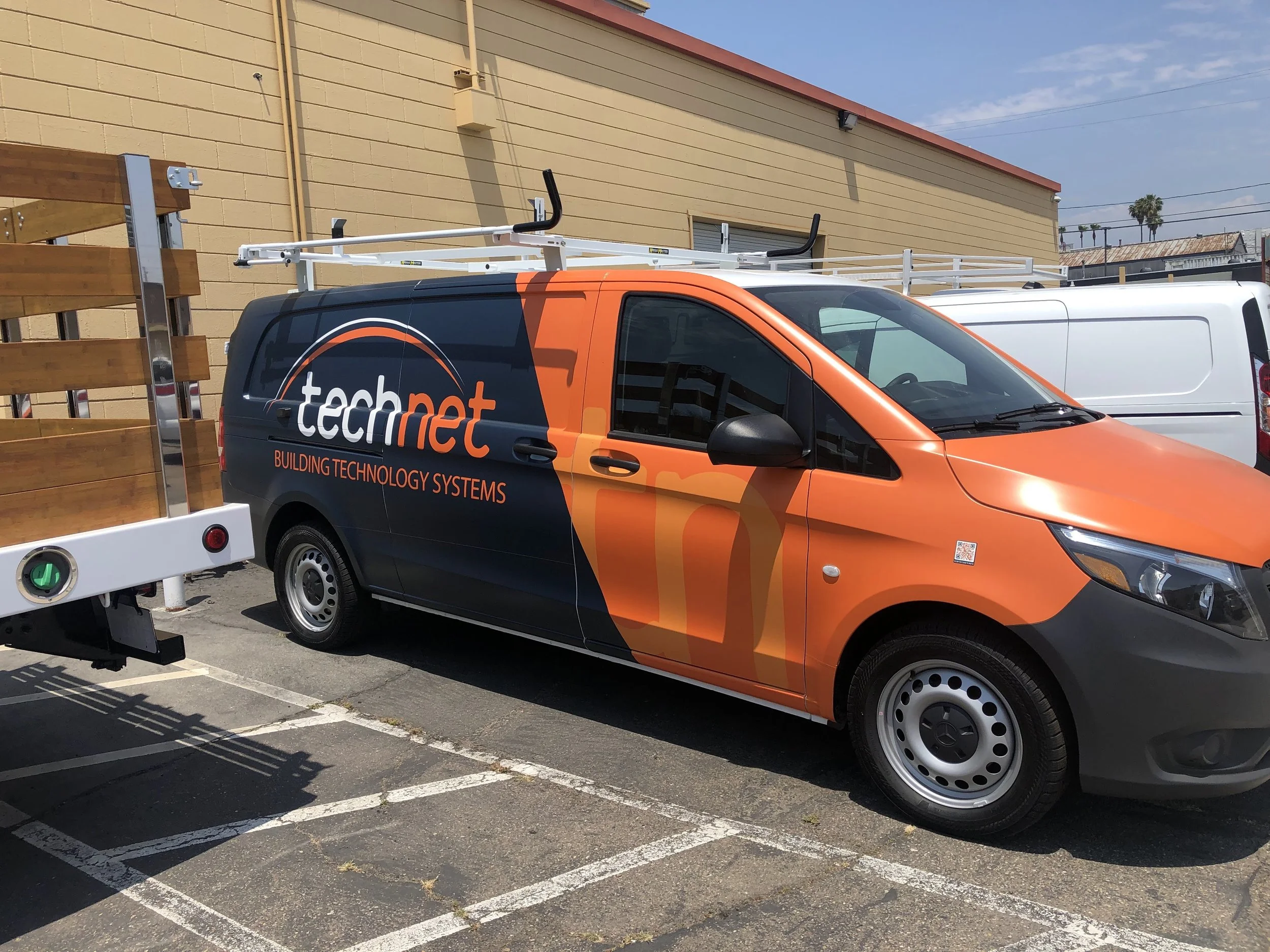 A black and orange work van with the logo and name 'tech net' and the slogan 'Building Technology Systems' parked in an outdoor parking lot next to a beige building and a white van.