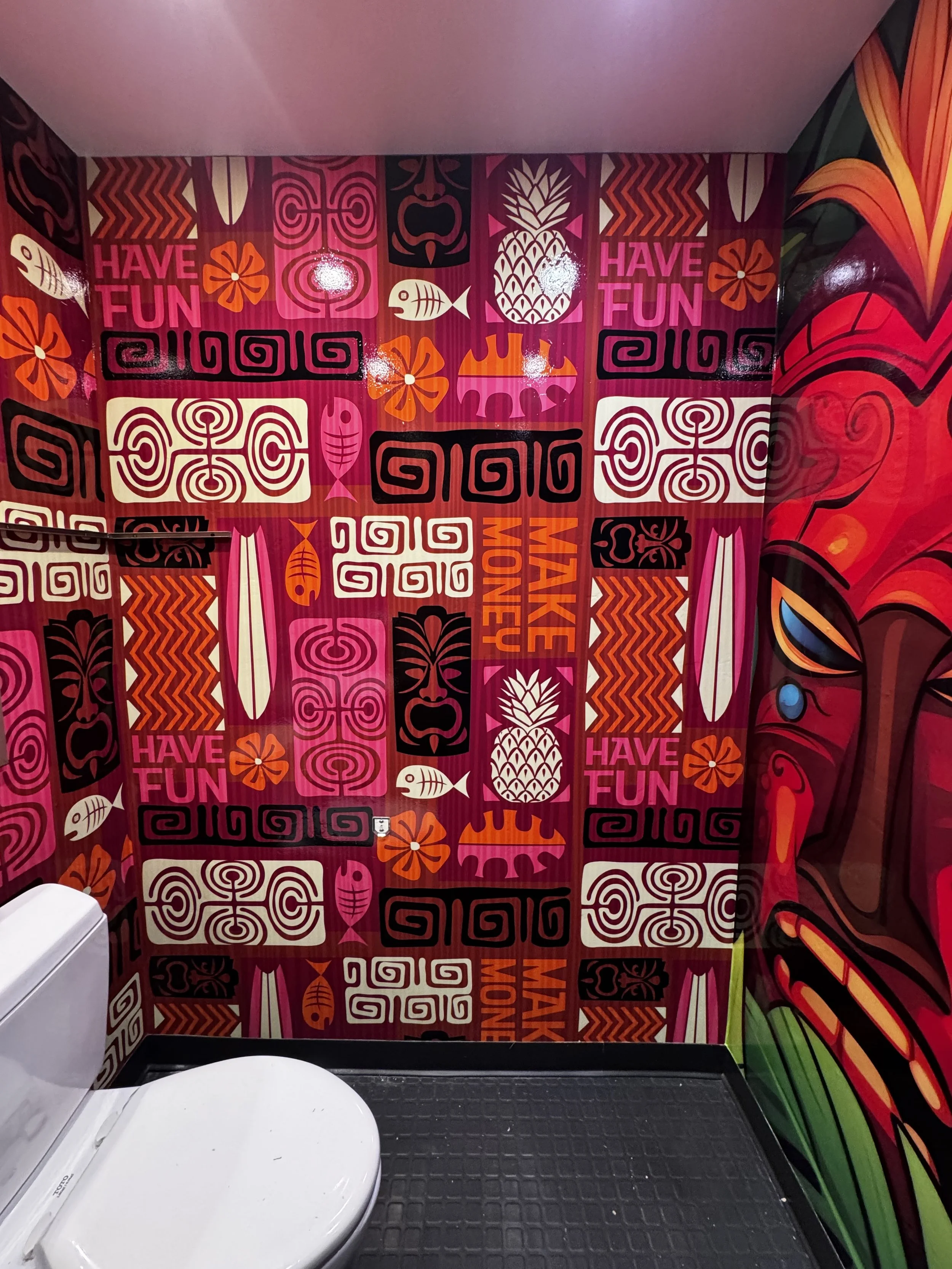 Colorful tropical-themed bathroom with a mural featuring phrases like 'Have Fun', 'Make Money', and tropical images such as pineapples, fish, and flowers, with a white toilet and black rubber mat on the floor.