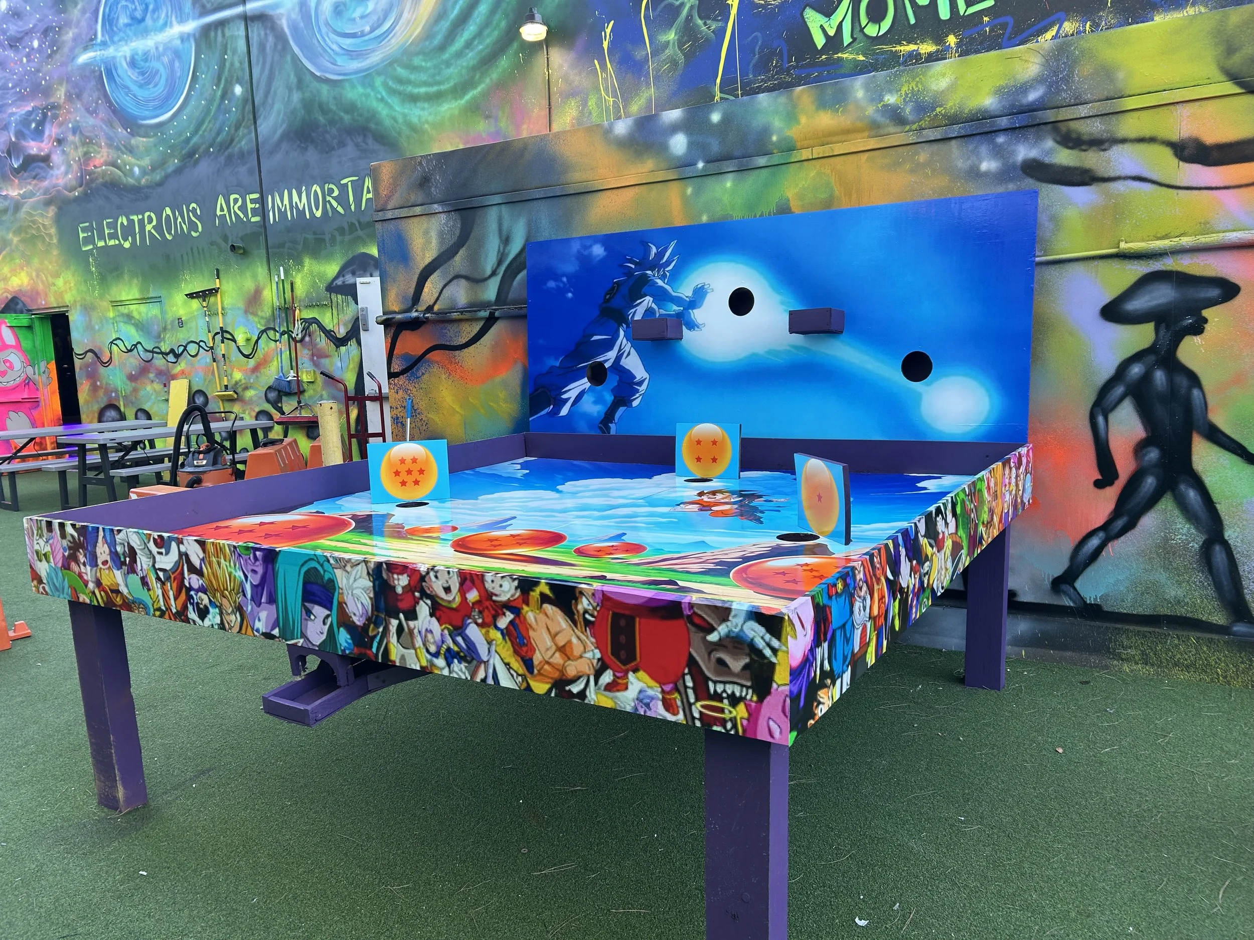 Colorful graffiti-covered air hockey table with Dragon Ball Z theme, featuring anime characters and symbols, set against a vibrant mural wall with space-themed and abstract art.