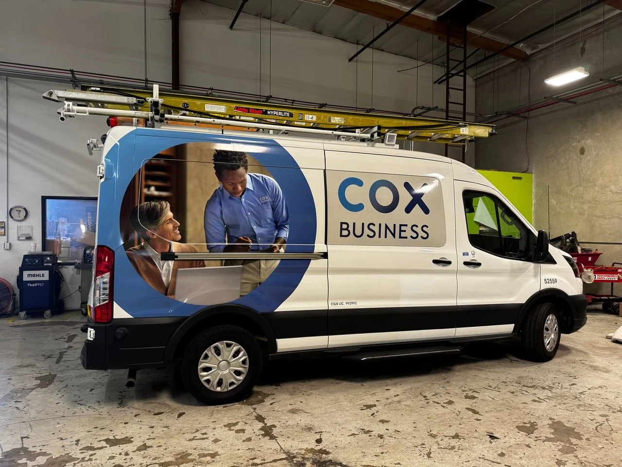 A commercial van with COX BUSINESS logo and branding, featuring a large graphic of two people, a man and a woman, engaging in a discussion inside a circle design on the side, inside a garage or workshop.