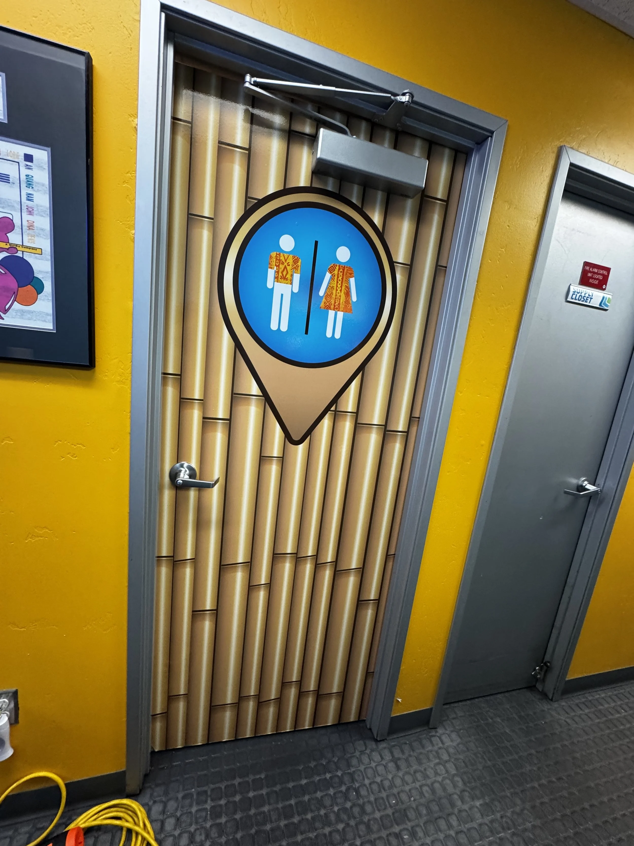 Restroom door with a sign of a man and woman in Hawaiian shirts and dresses, indicating a bathroom for both genders, in a tropical-themed setting.