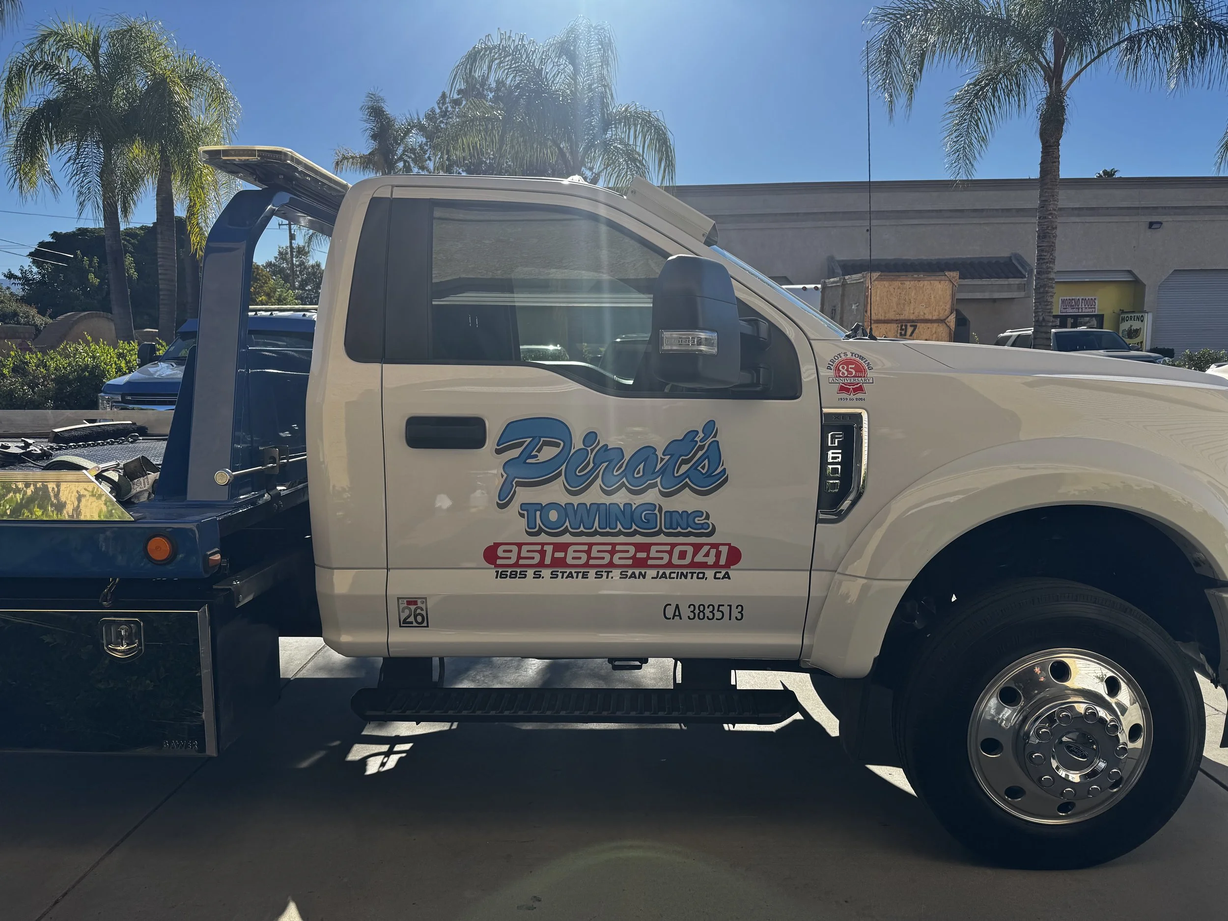 A white tow truck with the company name Piotr's Towing Inc. and contact information on the side, parked outdoors under palm trees.