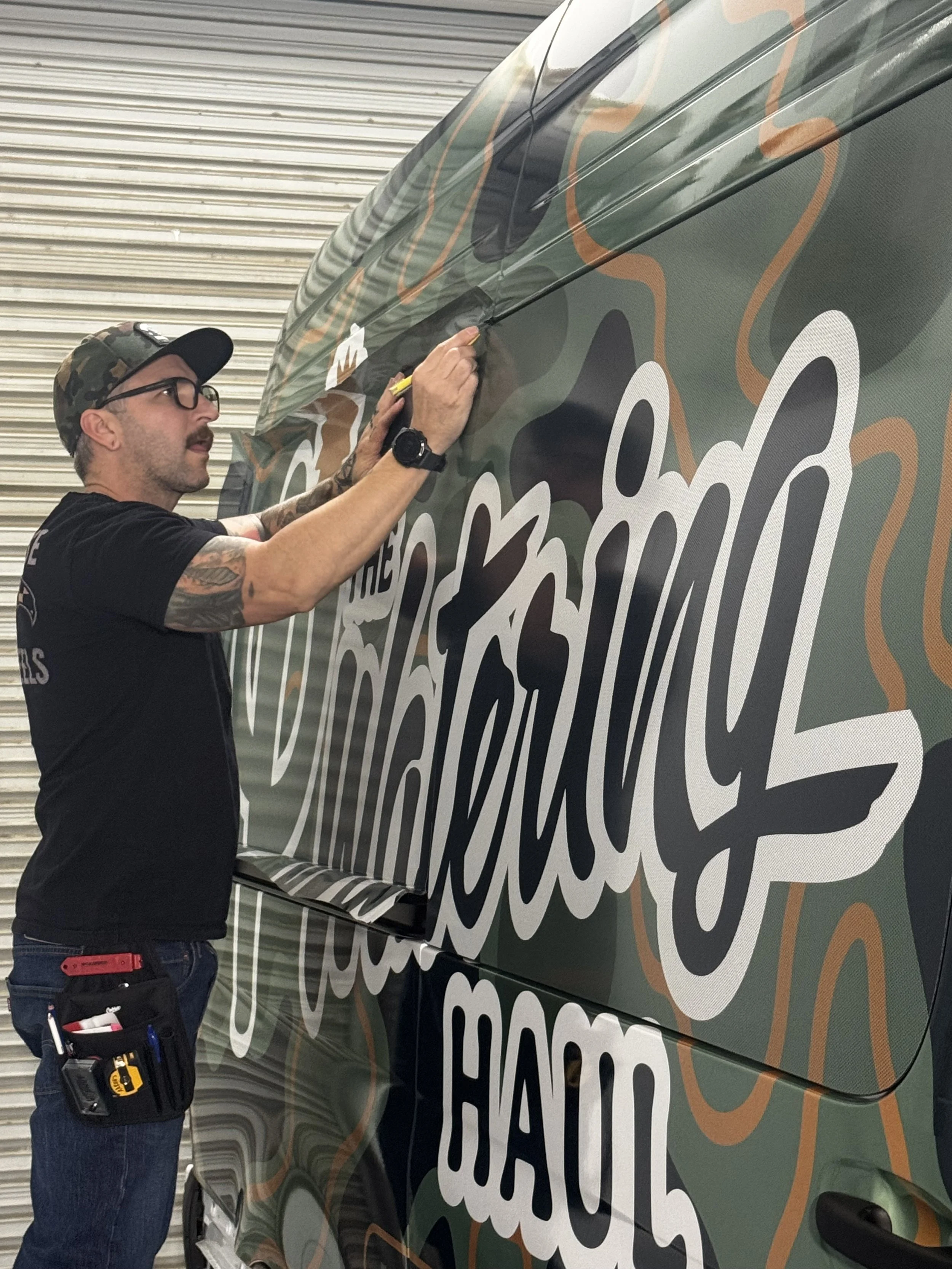 A man with glasses and tattoos is applying a decal to a vehicle with camouflage design.