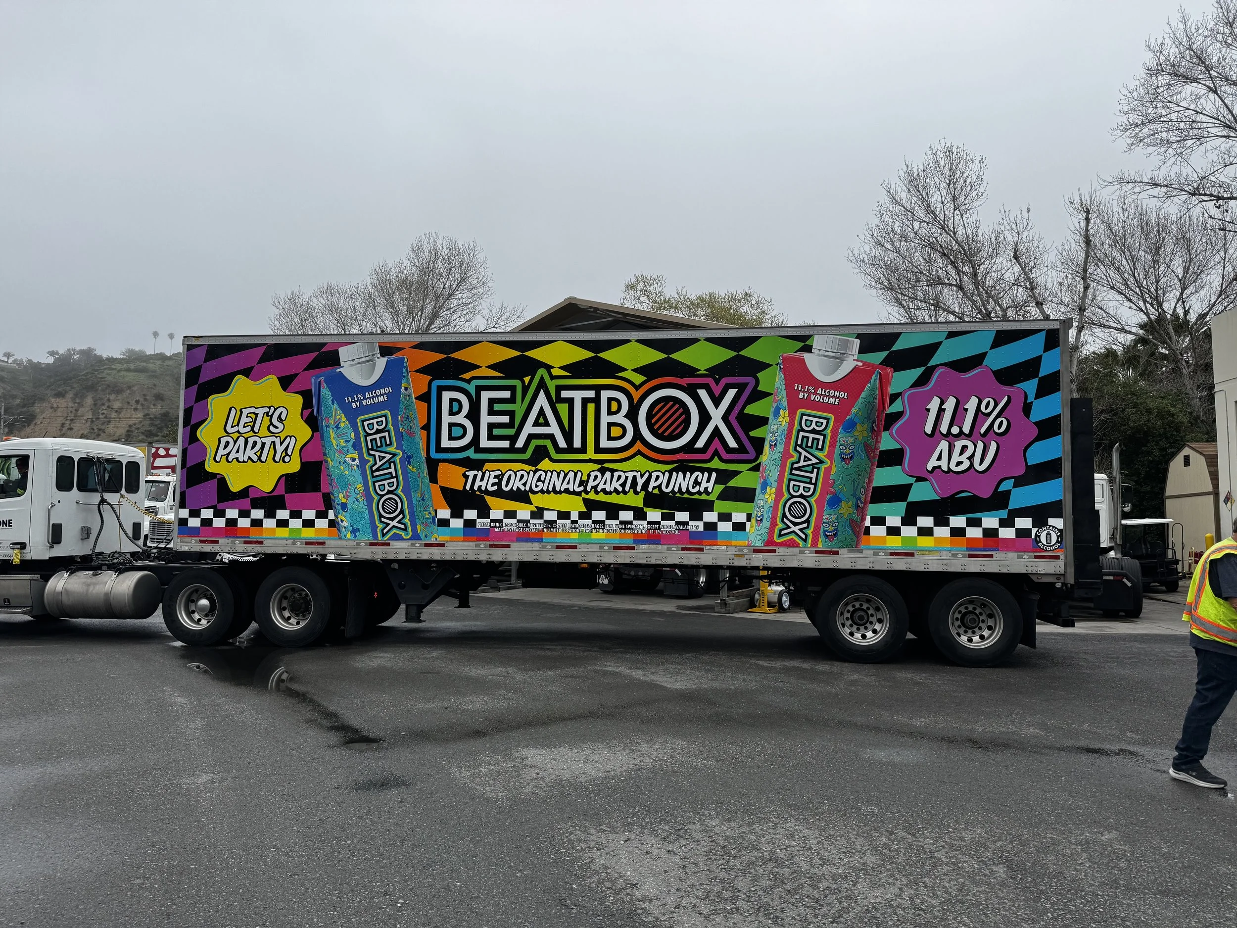 A truck with colorful graphics advertising BeatBox, a party drink, on its side, with text saying "Let's Party!" and "The Original Party Punch," featuring bottles labeled 11.1% alcohol and 11.1% ABV. There are trees and overcast sky in the background,