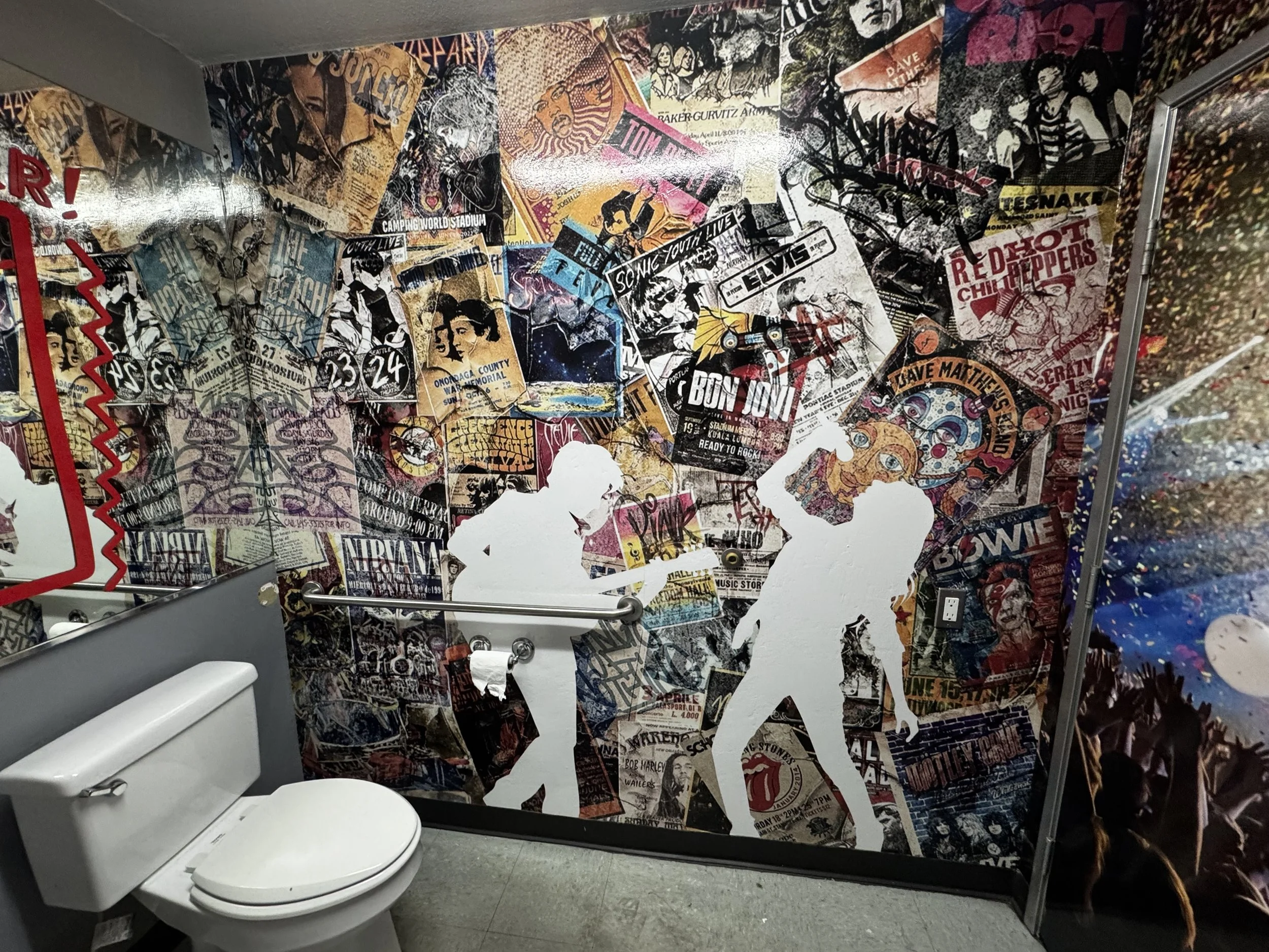 A decorative wall in a bathroom covered with a collage of music posters and album covers, featuring silhouettes of two people walking, a toilet, and part of a mirror on the left side.