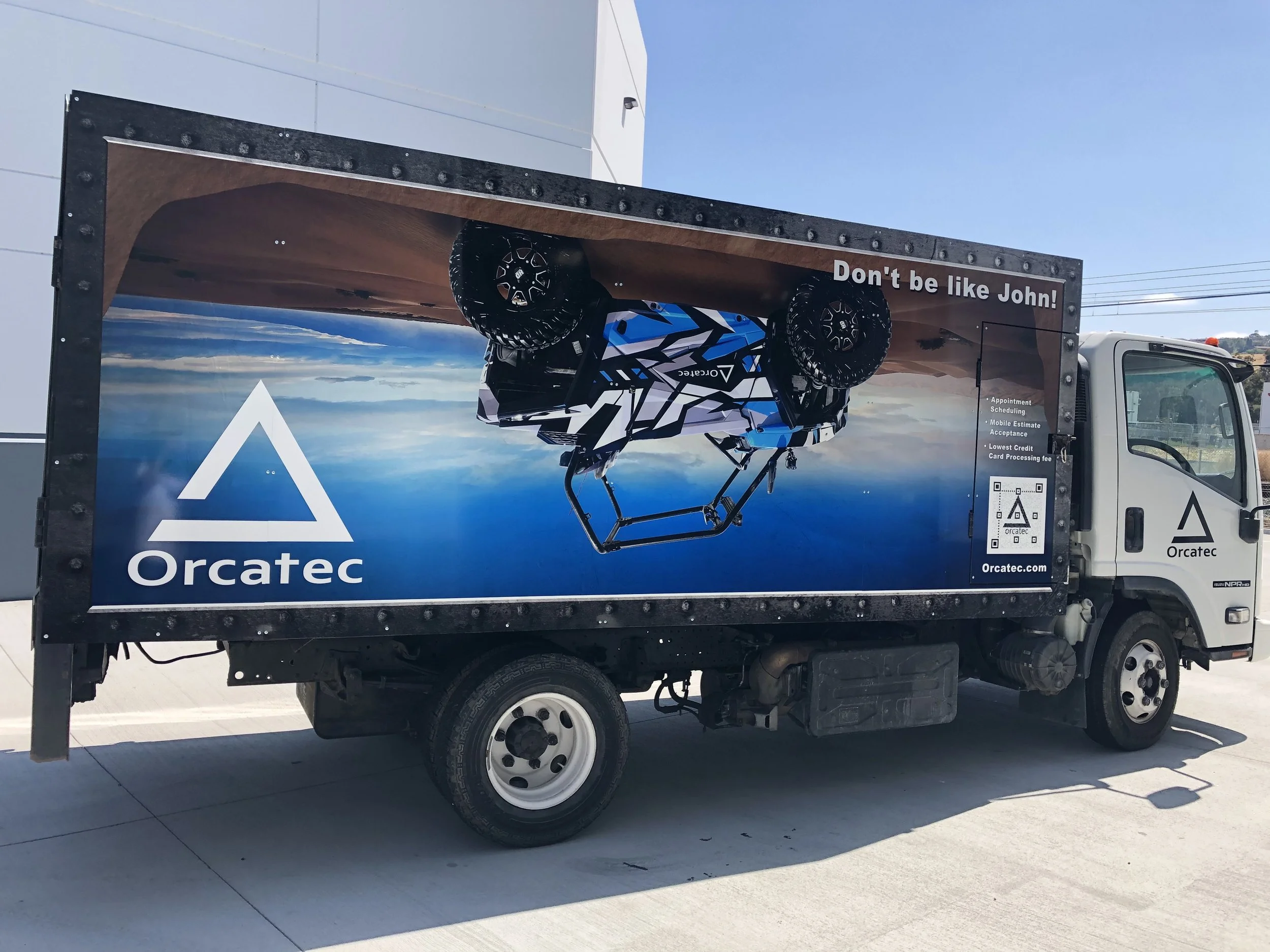 A delivery truck with a large advertisement on its side showcases an aerial view of a helicopter with a blue and black color scheme, and the text "Don't be like John!" is visible.