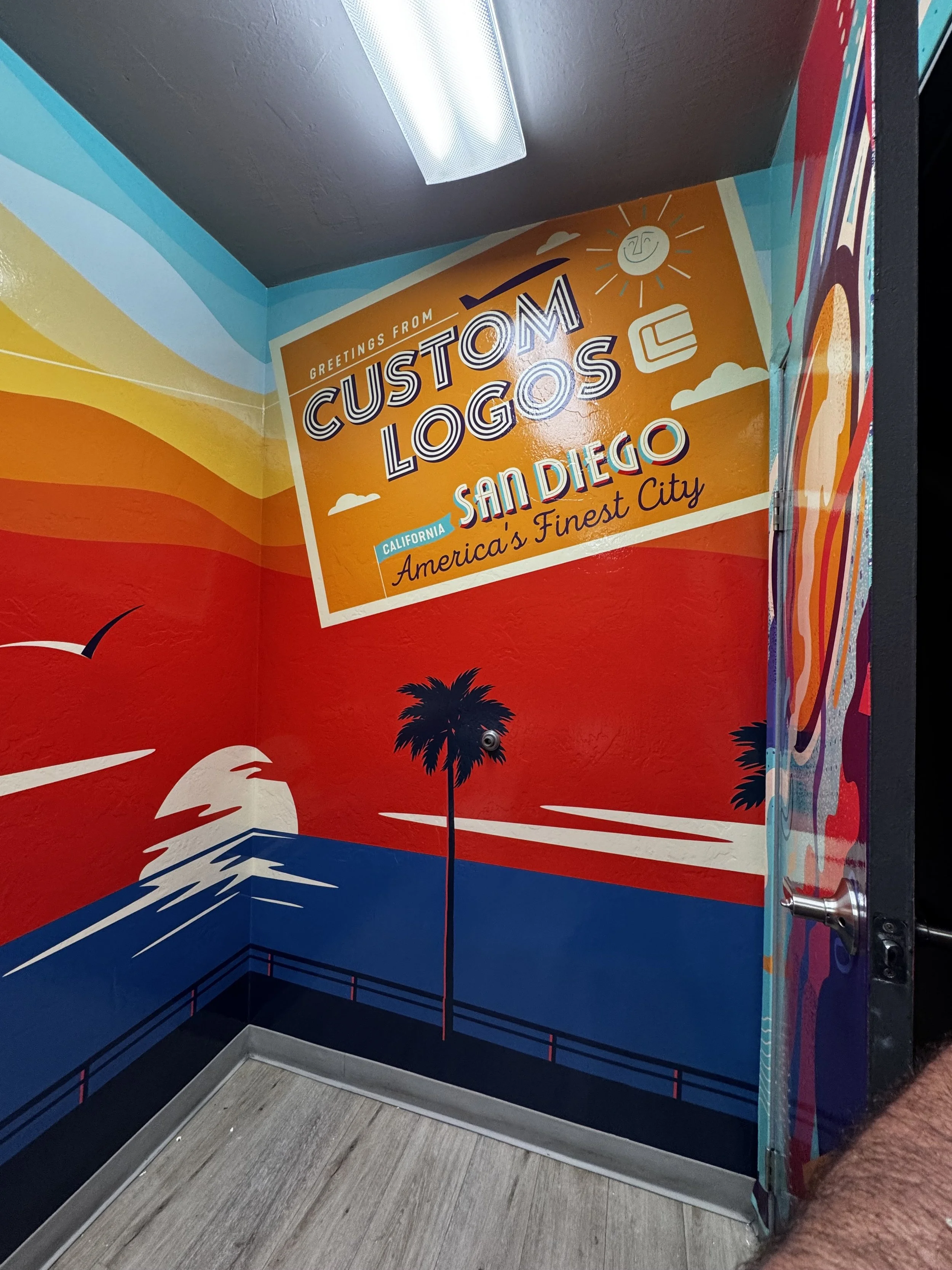 Colorful mural inside a phone booth featuring a sign that reads 'Greetings from Custom Logos San Diego, America's Finest City'. The mural depicts a sunset over the ocean with a palm tree and seagulls, and the overall style is vibrant and retro.