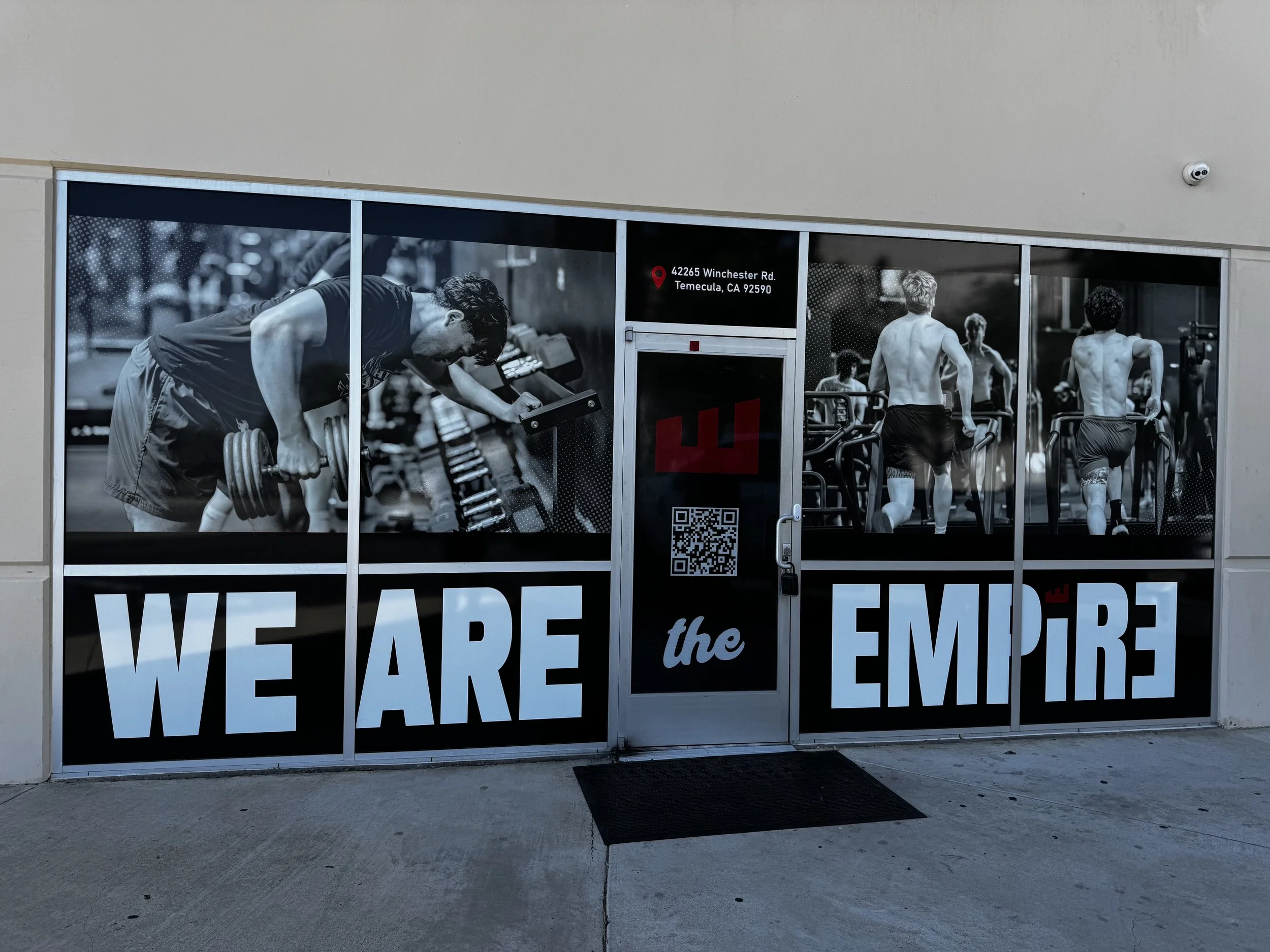 Black and white photo of people working out at a gym, with large text saying "WE ARE the EMPIRE" on the window.