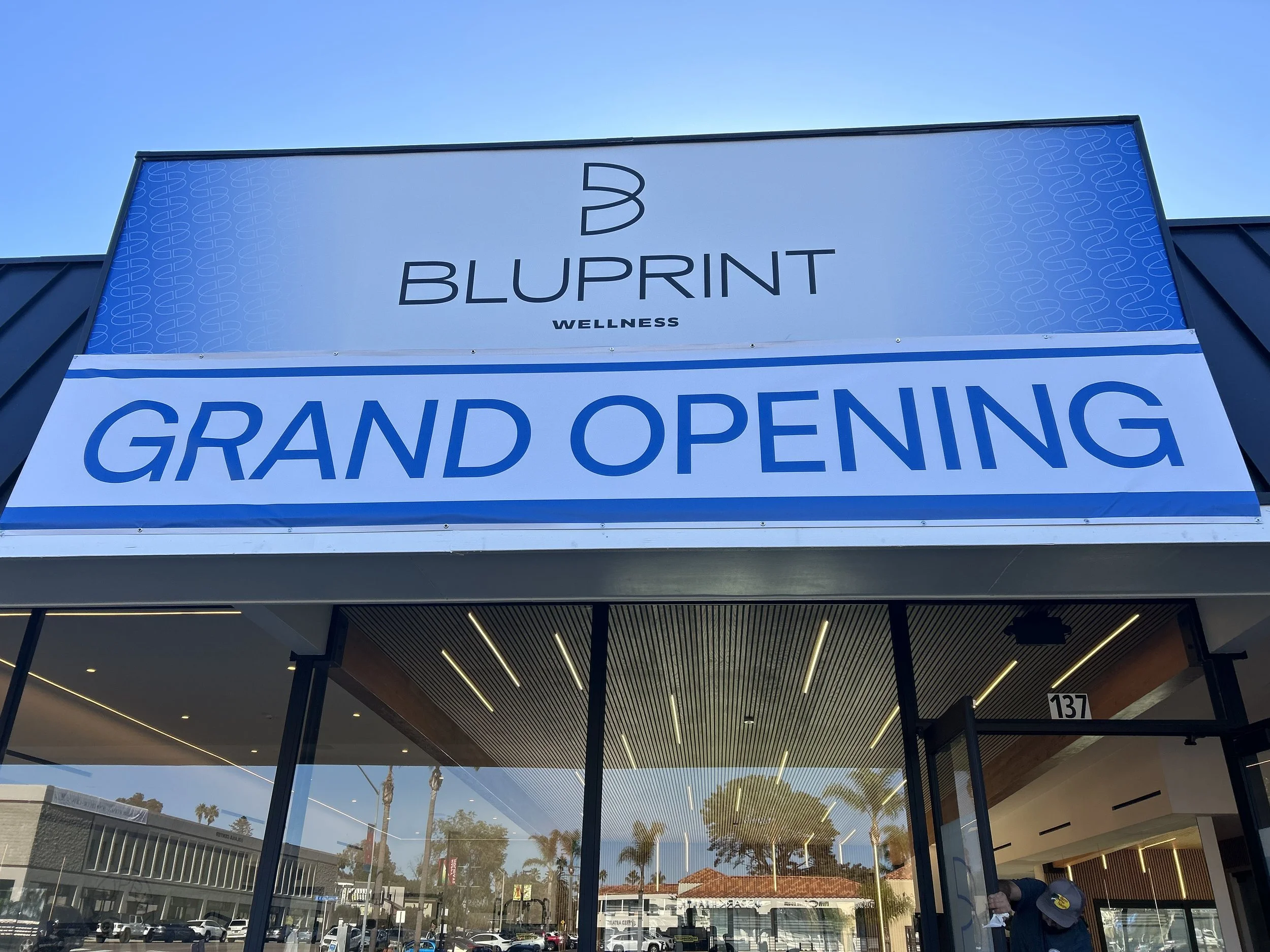 Storefront with large sign announcing 'Grand Opening' at Bluprint Wellness, with glass doors and a view of a shopping area outside with palm trees.