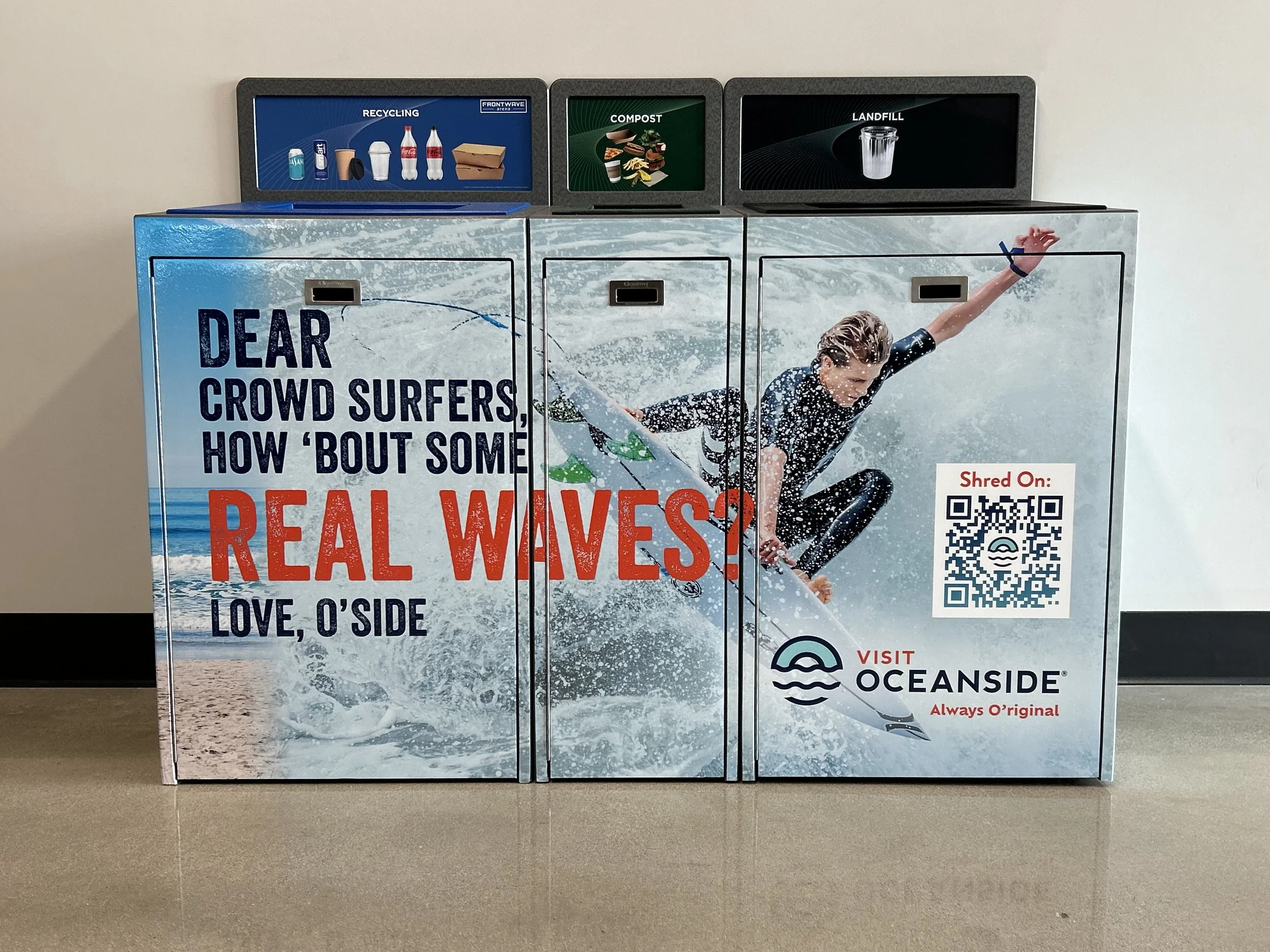 A vending machine decorated with a large surfing-themed graphic, featuring a female surfer on a wave, and promotional text encouraging surfing and visiting Oceanside, with recycling, compost, and landfill labels on top.
