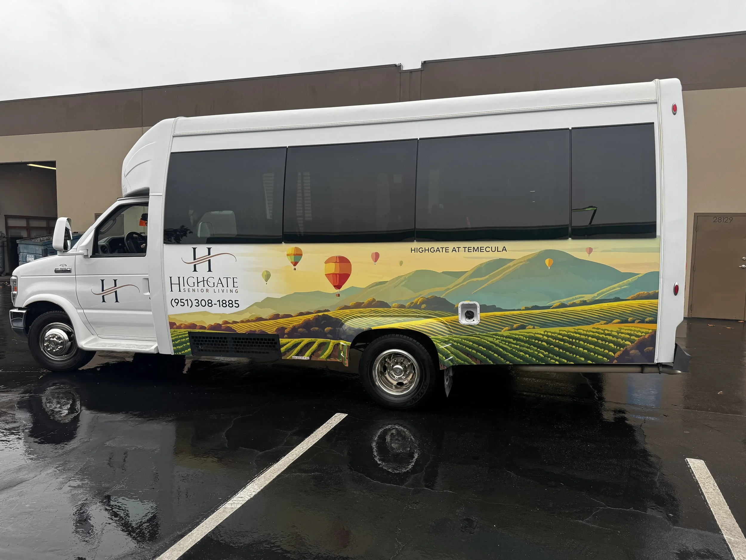 A white paratransit shuttle bus with a landscape mural of green hills and fields, hot air balloons, and a mountain range on its side, parked in a wet parking lot near a beige building.