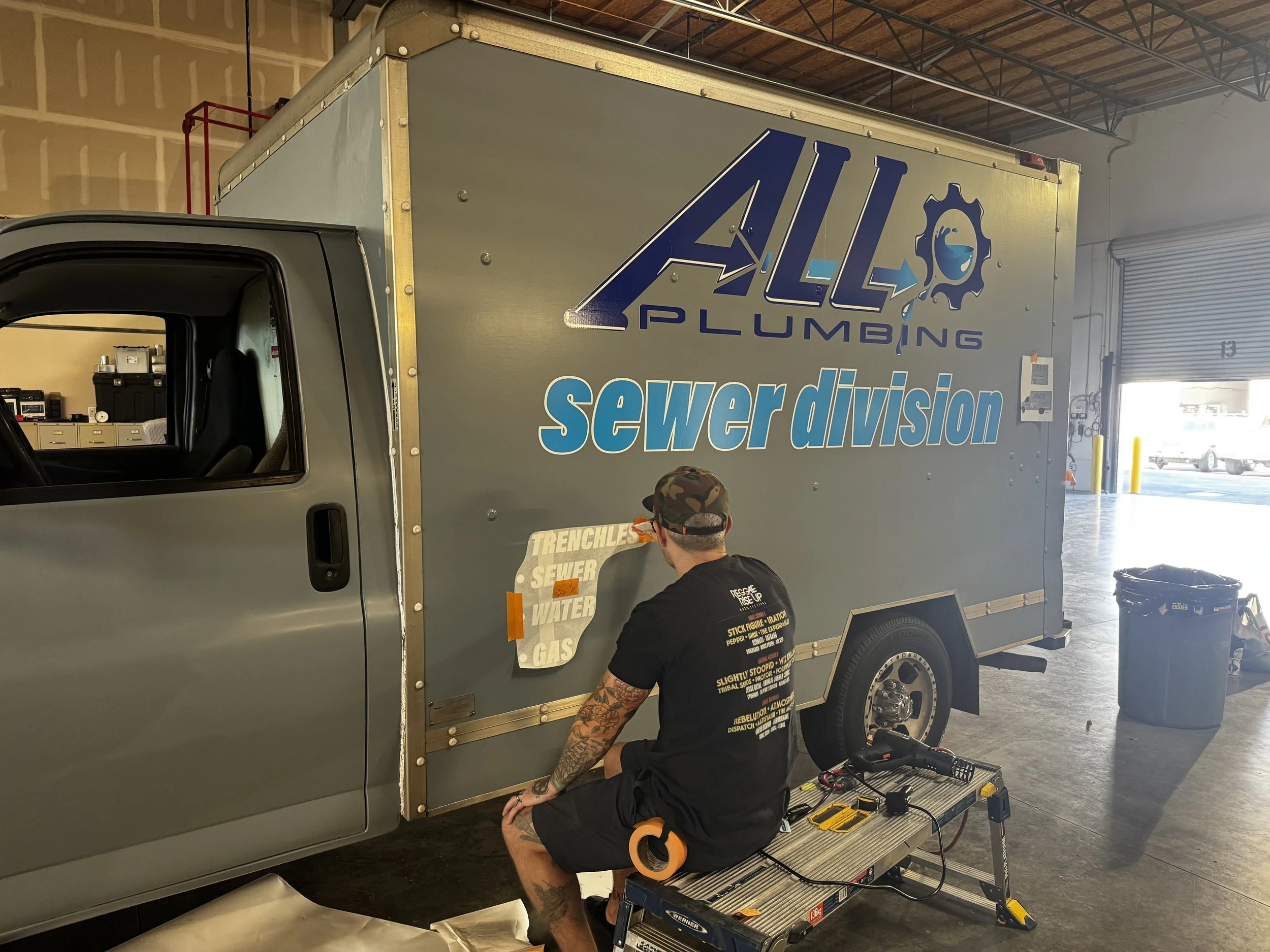A man sitting on a workbench welding or repairing a commercial plumbing truck inside a garage. The truck has large decals reading "All Plumbing" and "sewer division." The man is wearing a black T-shirt, shorts, and a camouflage cap and is focused on 