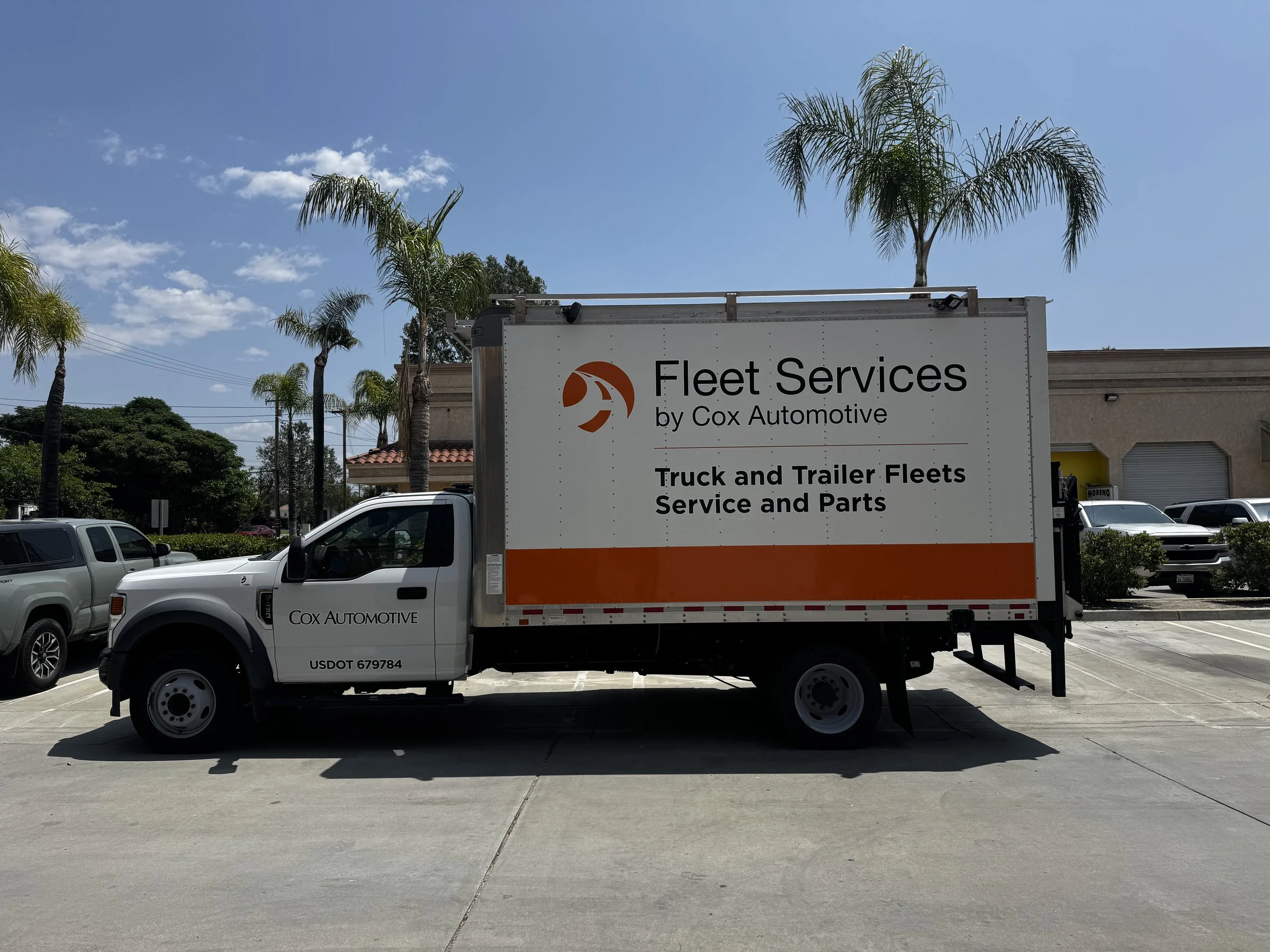A truck with the signage 'Fleet Services by Cox Automotive' parked in a lot with palm trees and a building in the background.