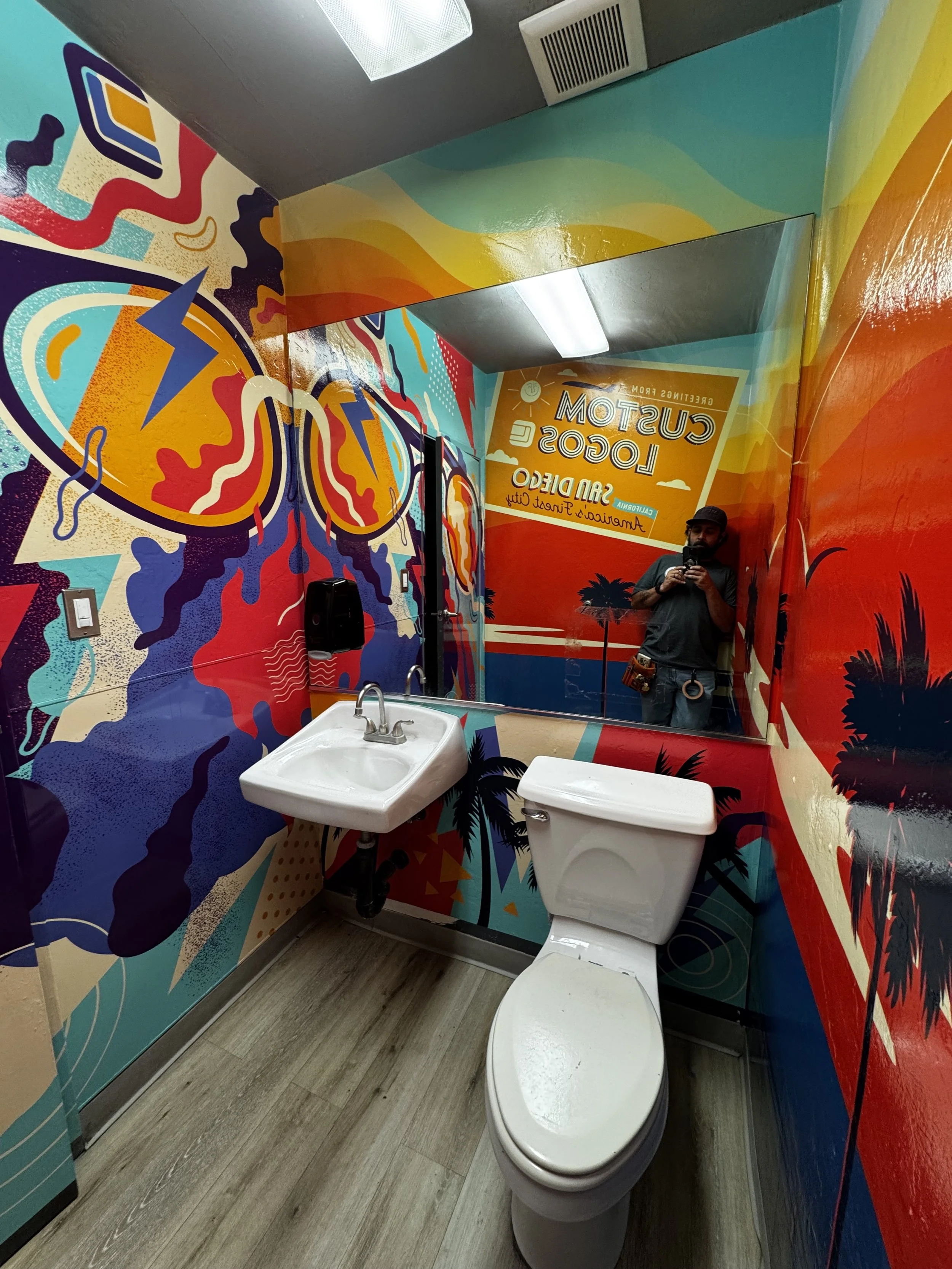 A vibrant tropical-themed bathroom with colorful wall murals depicting palm trees and abstract designs, featuring a small white sink, a white toilet, a large mirror, and a door. A person is taking a photo in the mirror.