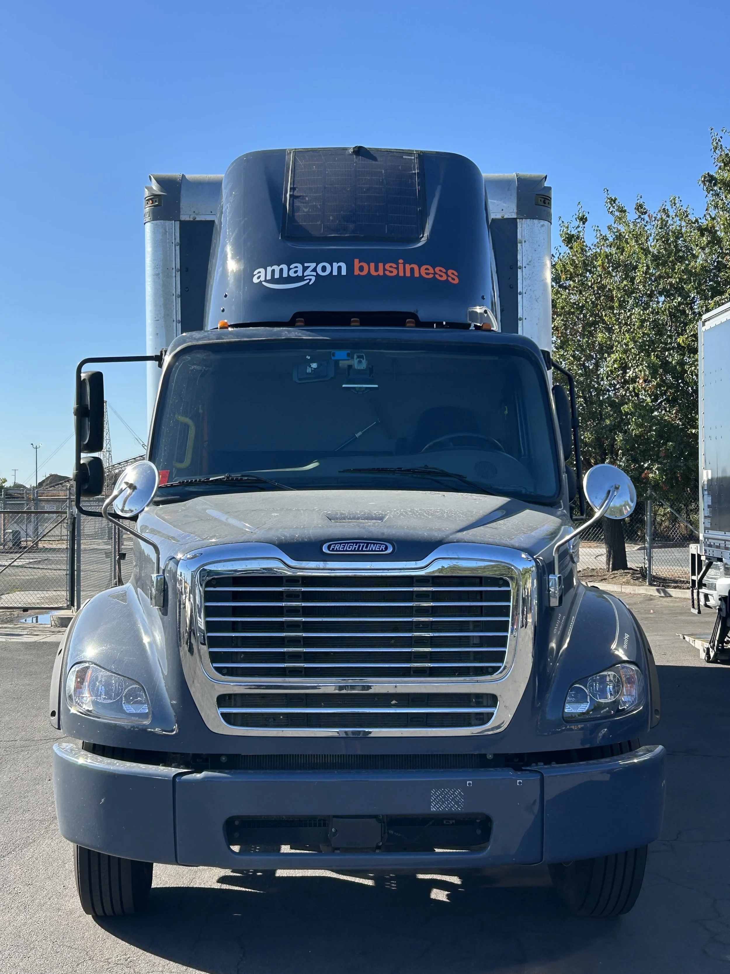 Front view of an Amazon Freightliner delivery truck parked outdoors under clear blue sky, with trees and a fence in the background.