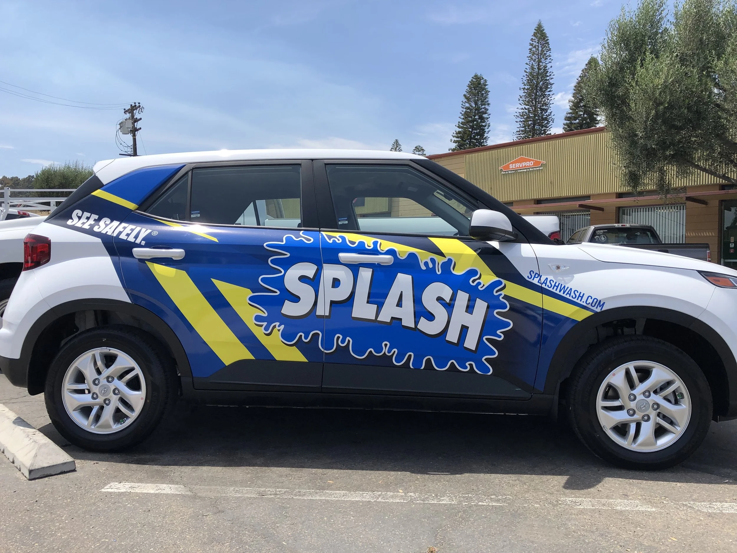 A vehicle with Splash branding, blue and yellow design, parked in a lot with a building and cars in the background.