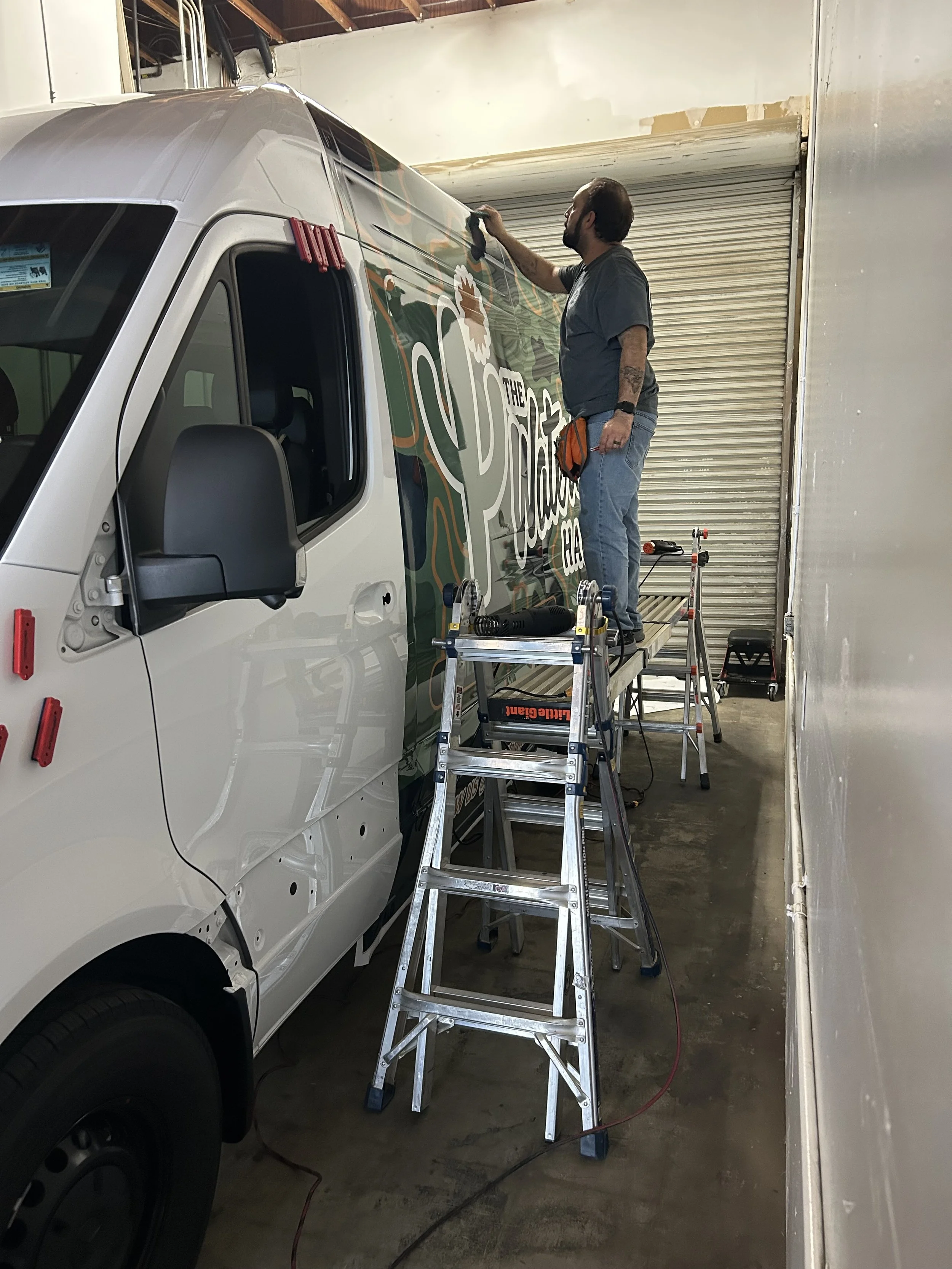 A man standing on a ladder painting a colorful mural on the side of a white delivery van inside a garage.