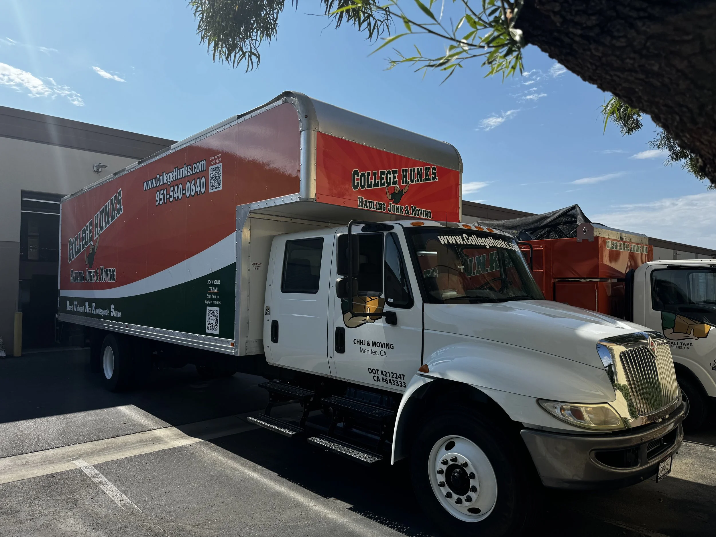 A moving truck with branding for College H.U.N.K.S. Hauling Junk & Moving parked in a parking lot under a tree.