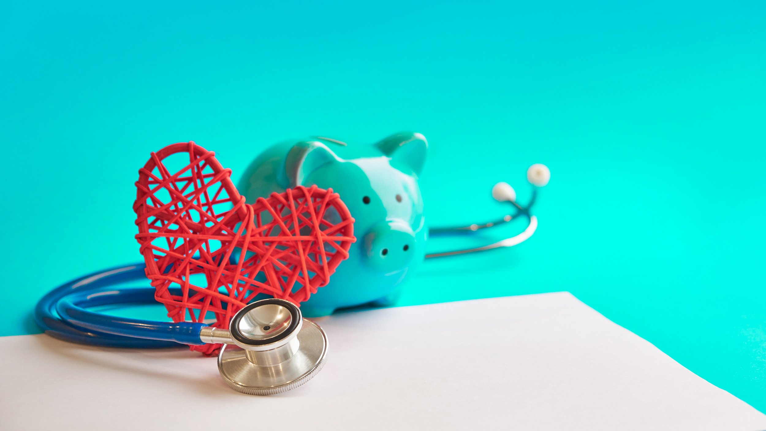 A stethoscope, a red woven heart decoration, a light blue piggy bank, on a white and turquoise background.