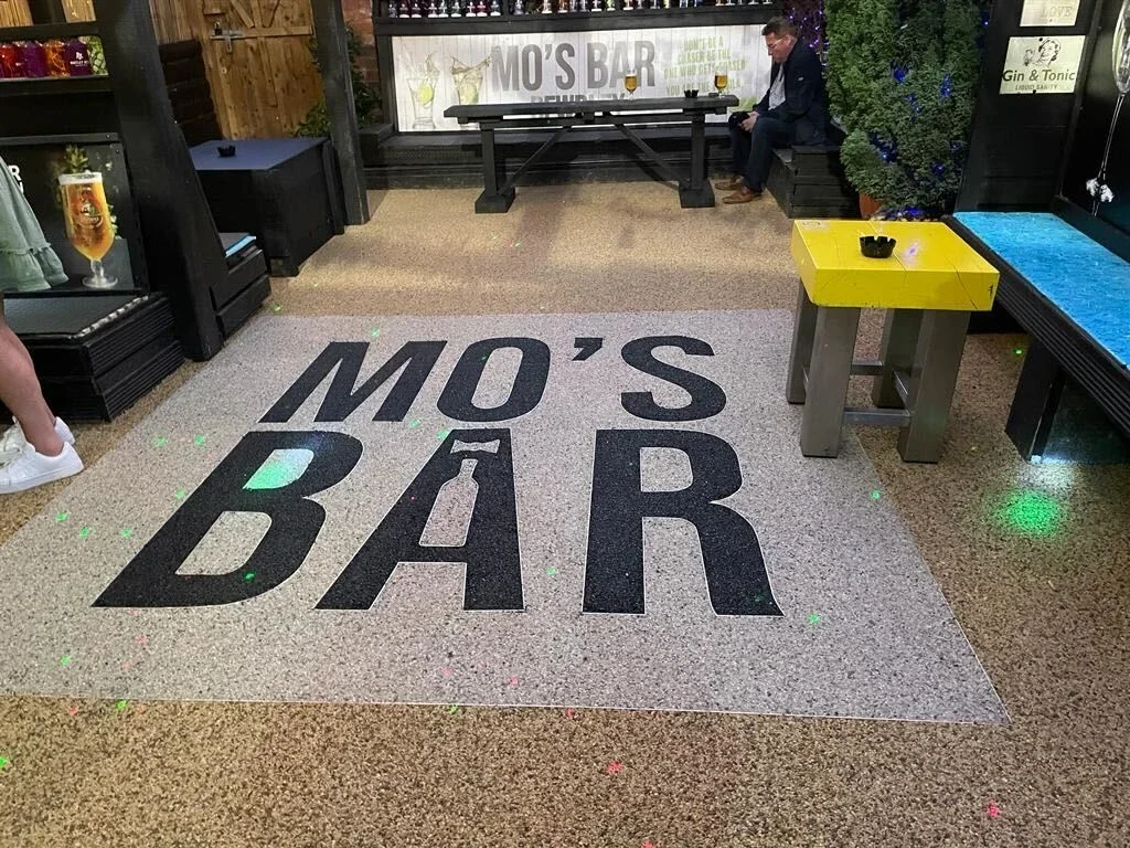 Interior of MO'S BAR in Perth featuring branded resin bound flooring with, rustic seating, yellow table with ashtray, blue-lit tree, and 'Gin & Tonic Lounge Bar' signage—casual nightlife venue with modern charm."