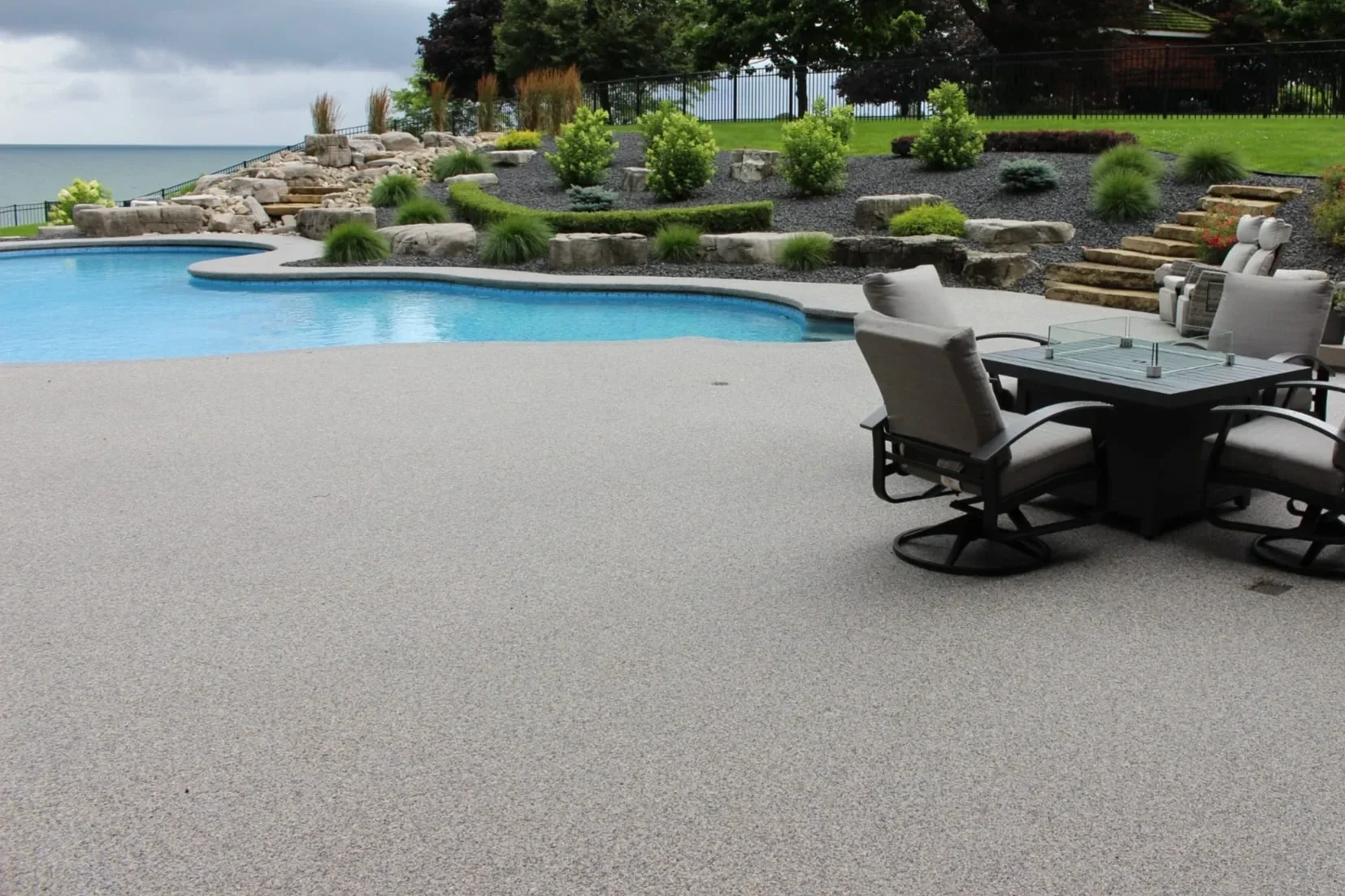 A Perth resin bound swimming pool surround , surrounded by a deck with outdoor chairs and a table, landscaped with rocks, shrubs, and trees.