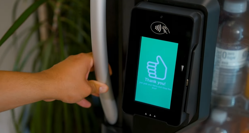 A person using a contactless payment terminal on a shopping cart handle with a screen showing a thumbs-up icon and 'Thank you!'