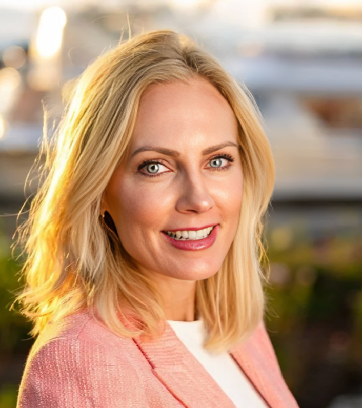 A blonde woman with blue eyes smiling outdoors, wearing a pink blazer and white top, with boats in the background.
