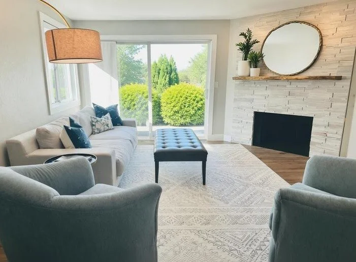 Living room with white sofa, blue pillows, two gray armchairs, a tufted black ottoman, a white brick fireplace with a wooden shelf, a round mirror, and a large window with greenery outside.