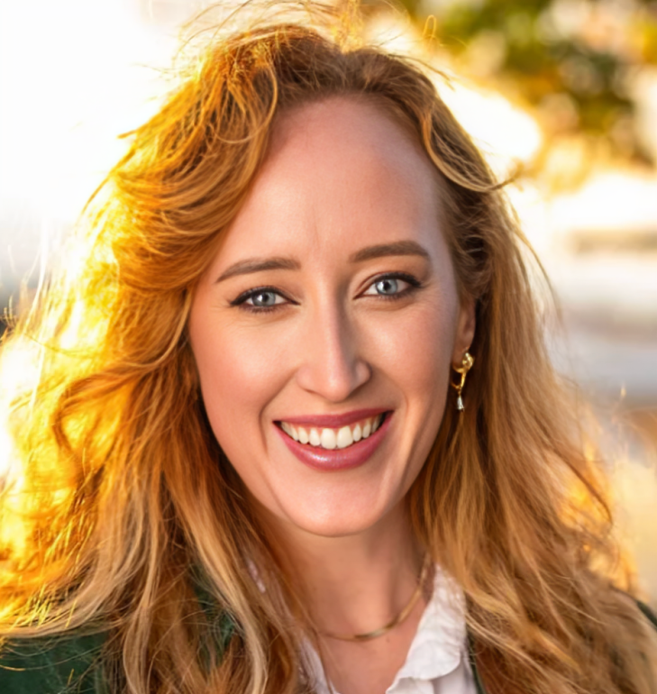 Smiling woman with long, wavy red hair outdoors during sunset, wearing gold earrings and a white top.