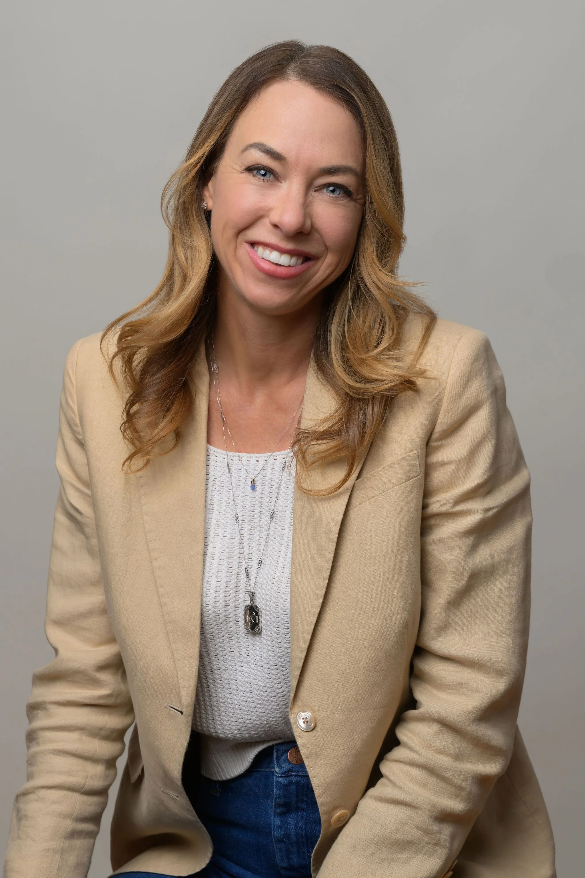 A smiling woman with wavy, shoulder-length blonde hair, wearing a beige blazer over a white textured top, with layered necklaces, against a plain gray background.