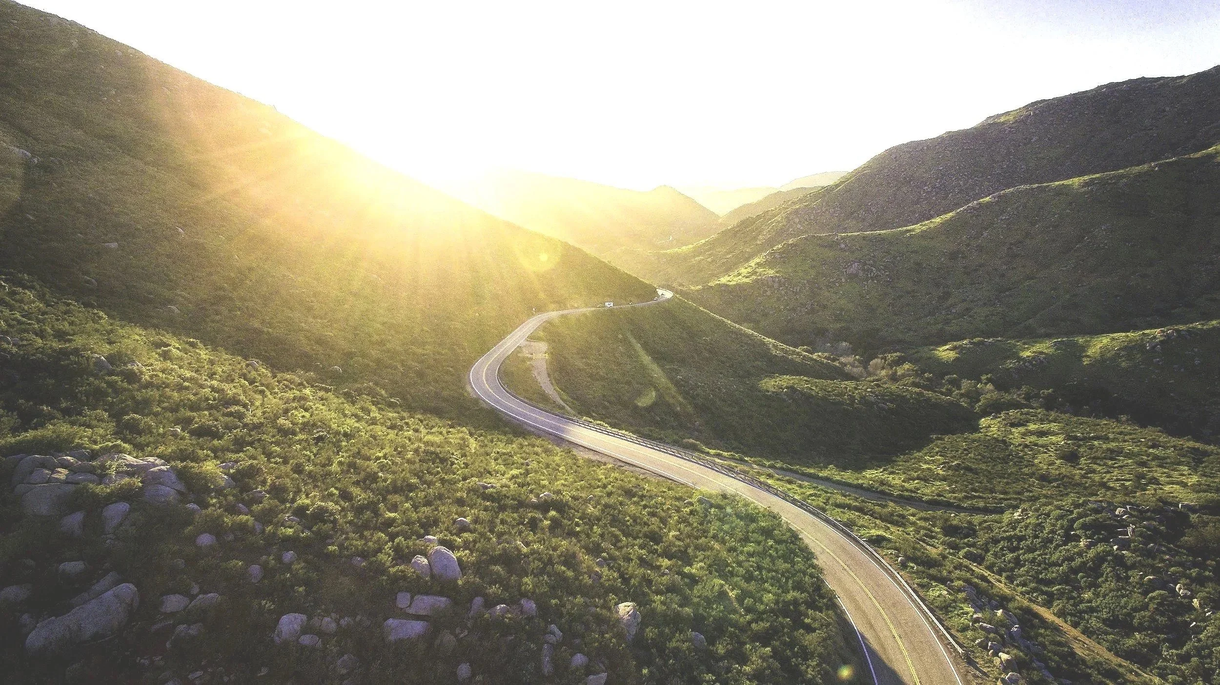 Winding mountain road with greenery on hillsides, shining sun over mountains in the distance.