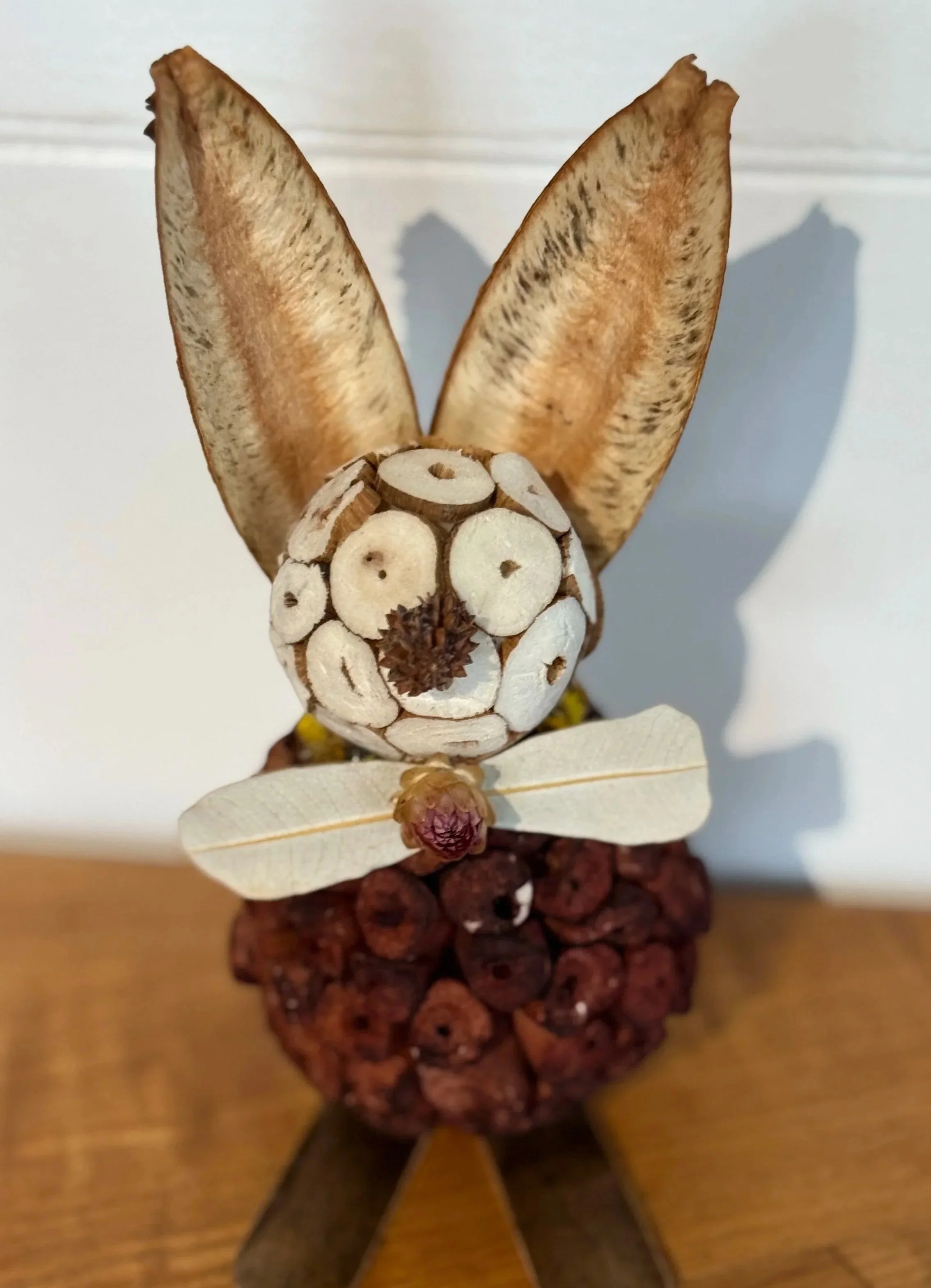 A bunny figure made of natural materials, with a body of small round objects, large dried leaves as ears, a face made of sliced small round items, a tiny cone for a nose, and wings resembling dragonfly wings, sitting on a wooden surface.