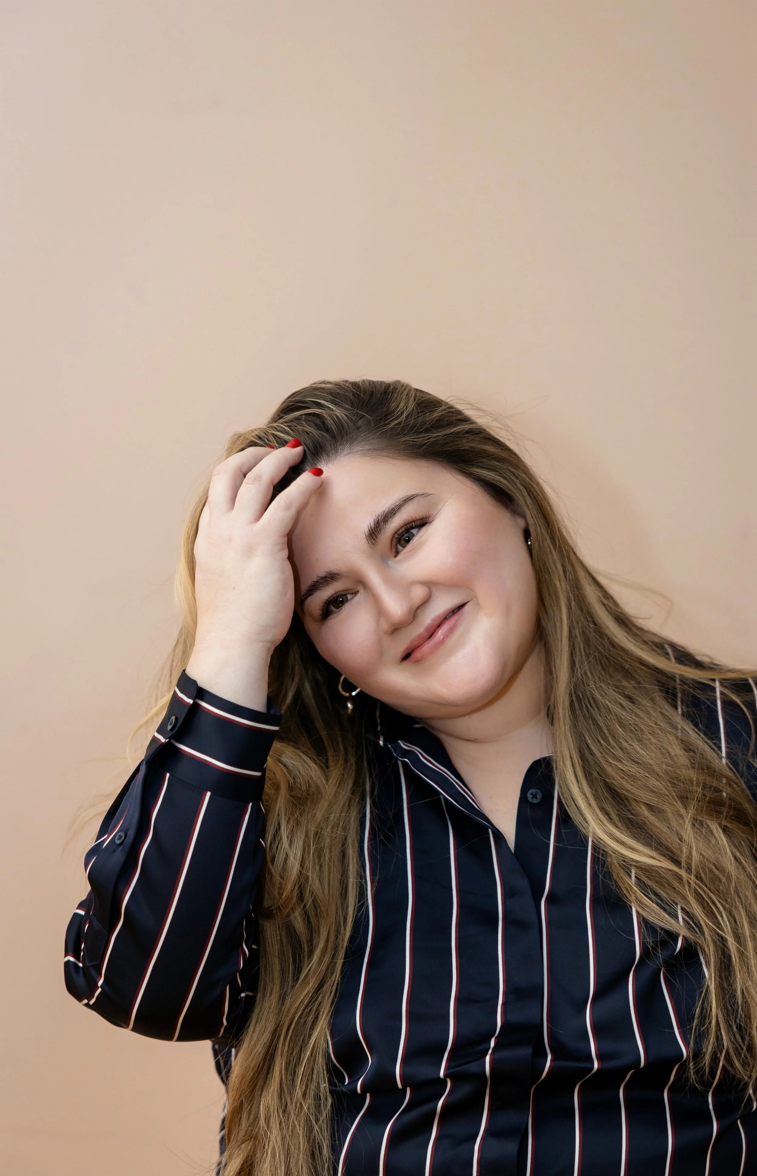 A smiling woman with long brown hair, wearing a navy blue striped shirt, resting her hand on her forehead with red painted nails, against a neutral beige background.