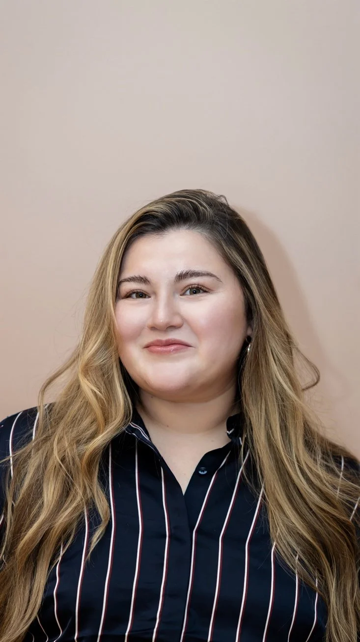 A young woman with long, wavy, light brown hair smiling at the camera with a plain beige background.