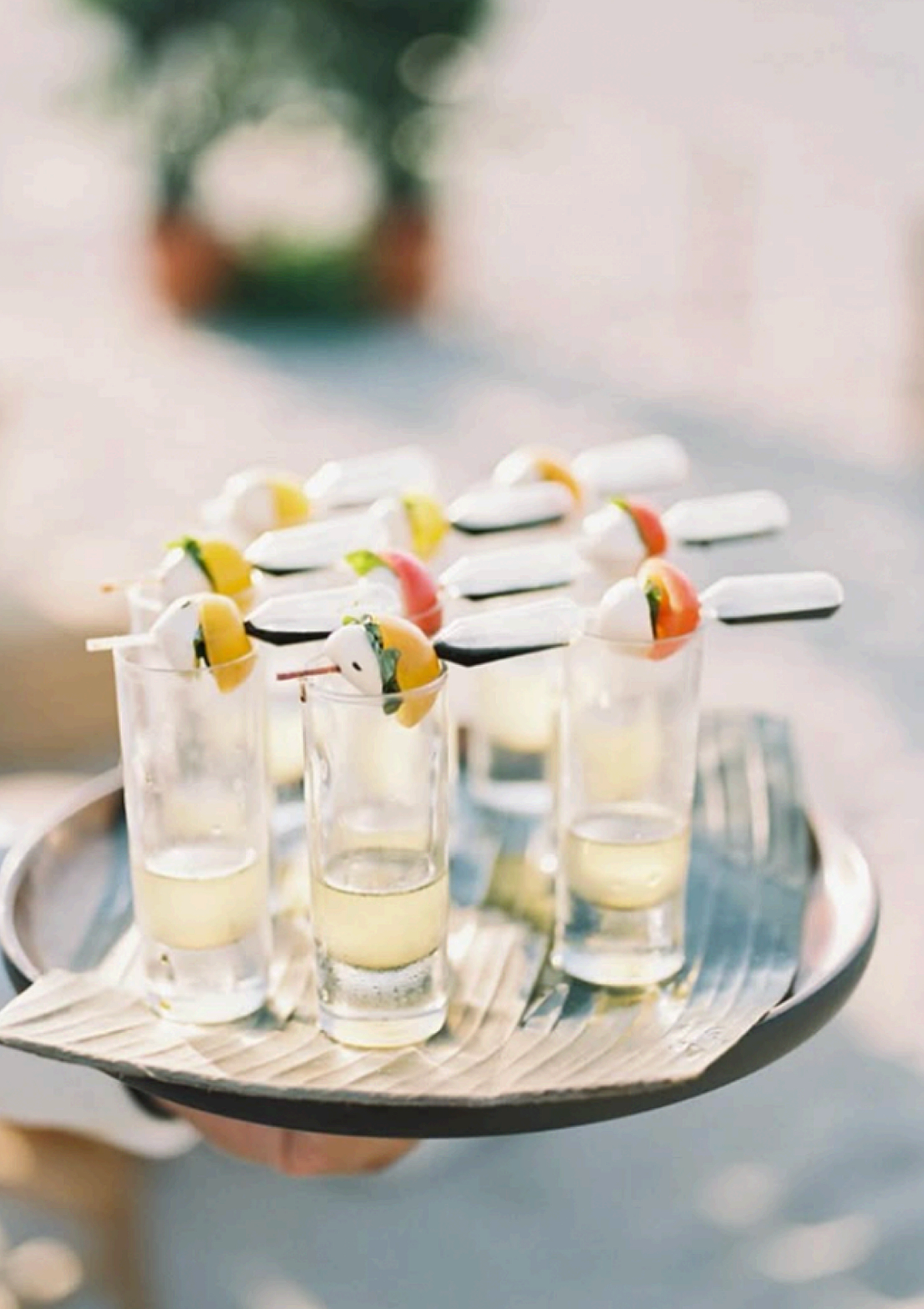 Shot of several small glasses filled with a yellow liquid, topped with colorful appetizers skewered on sticks, on a tray.