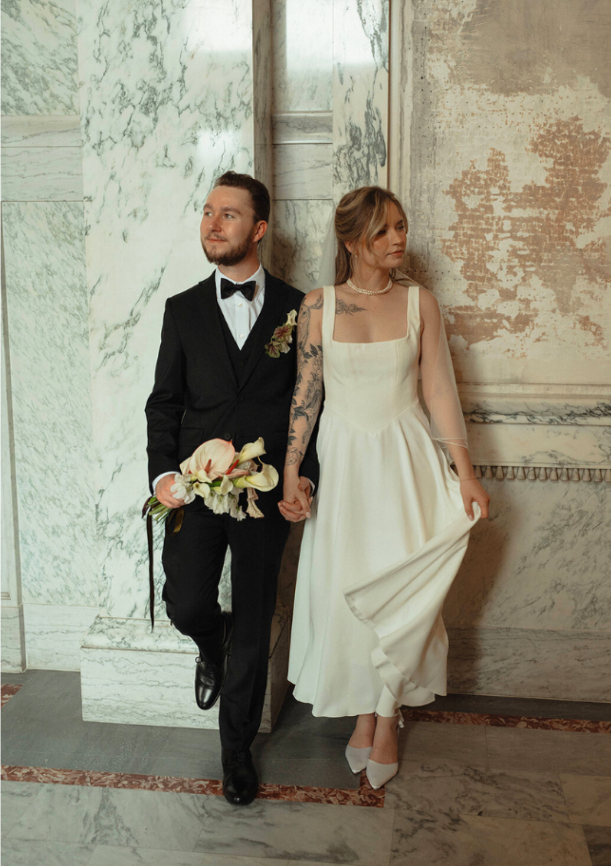 A newlywed couple stands against a marble wall, with the groom in a black tuxedo holding a bouquet and the bride in a cream dress holding her skirt, both looking to the side.