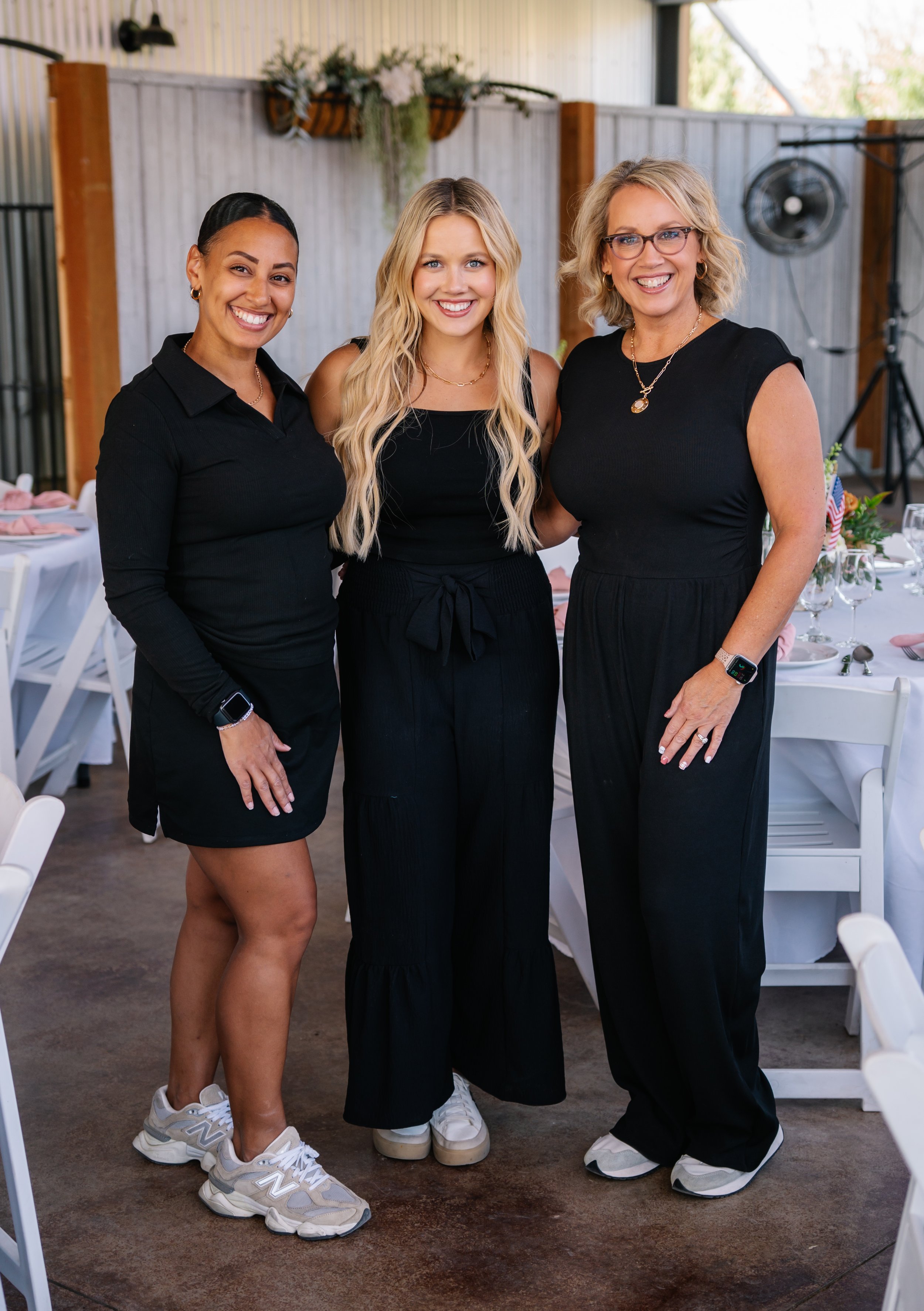 Three women standing together at a social event, dressed in black, smiling, with tables and chairs set for a gathering in the background.