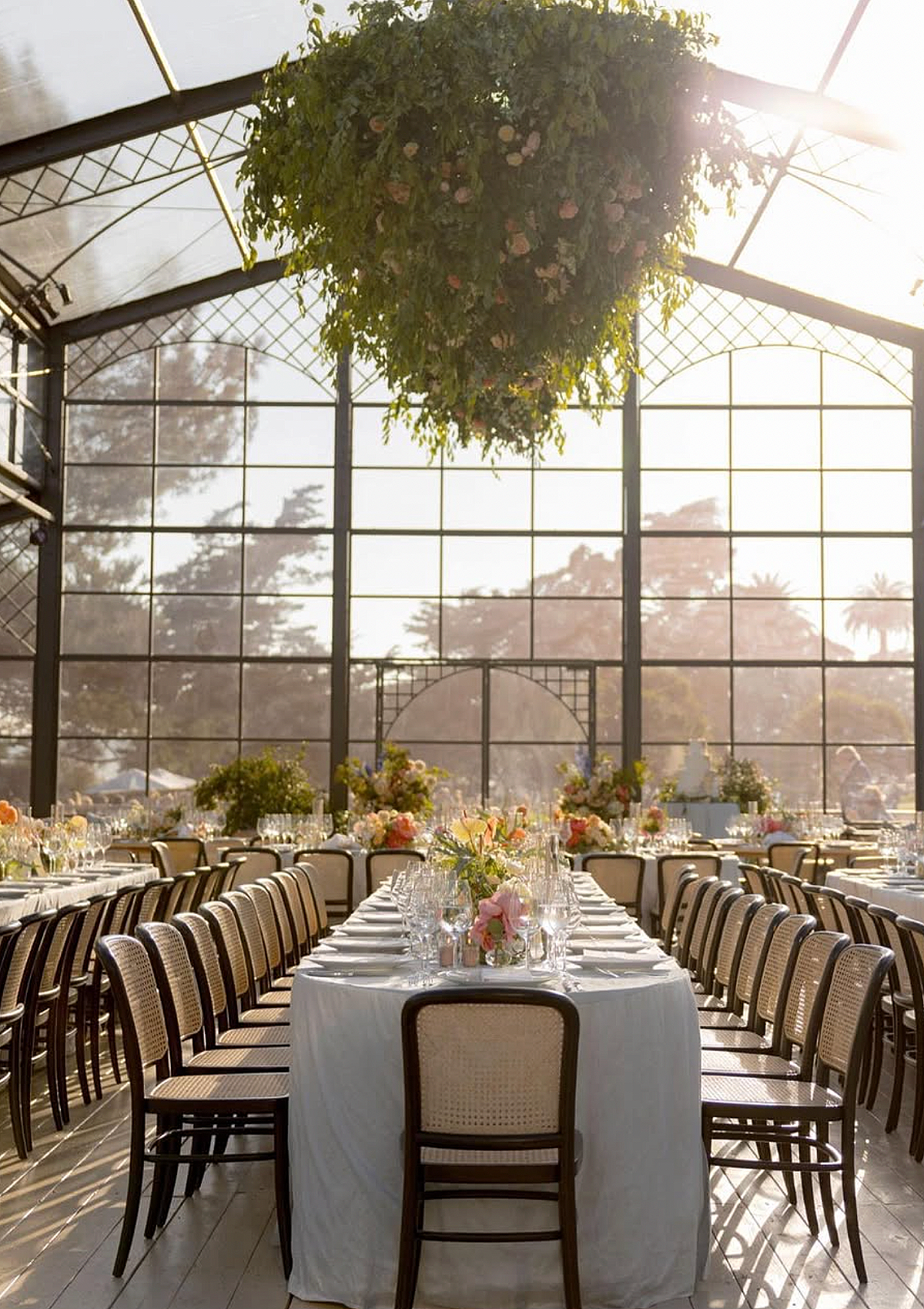 Elegant indoor wedding reception with a long table decorated with pink and white flowers, set with glassware and white tablecloths, in a glass conservatory filled with natural sunlight.