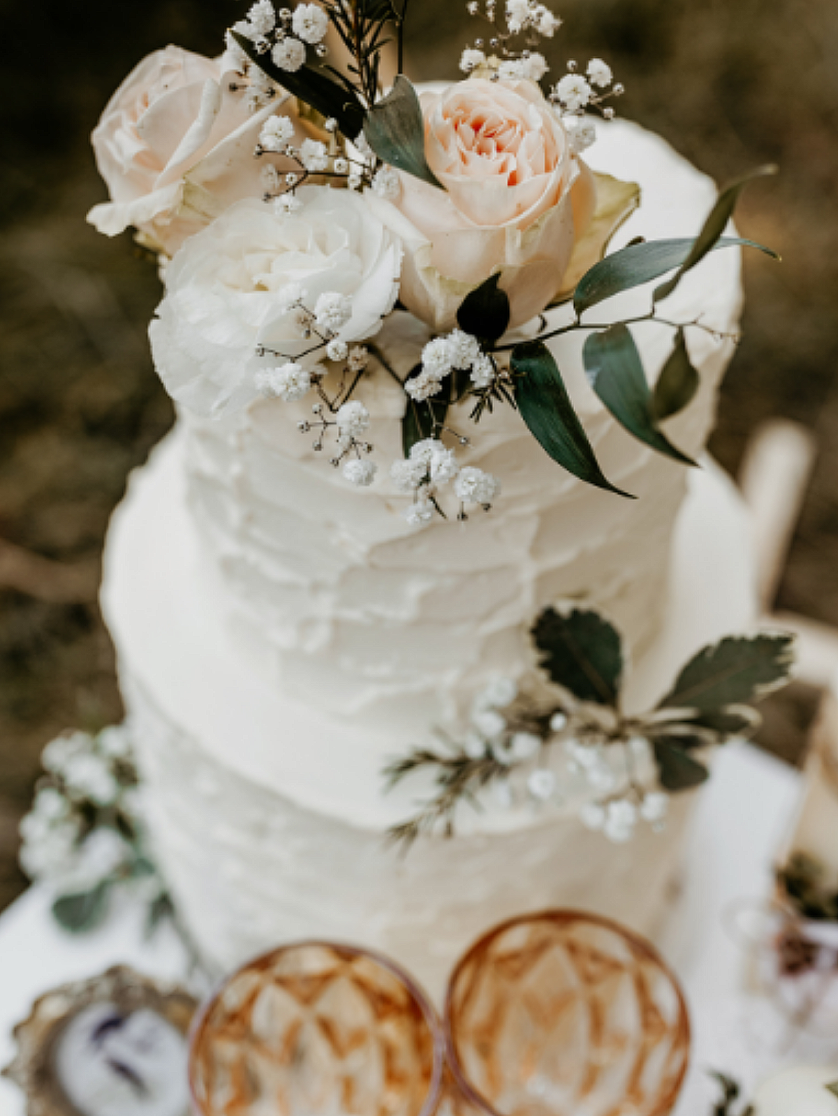 A three-tiered wedding cake decorated with white and blush roses, baby's breath, and green leaves, placed on a white table with orange and gold patterned glasses.