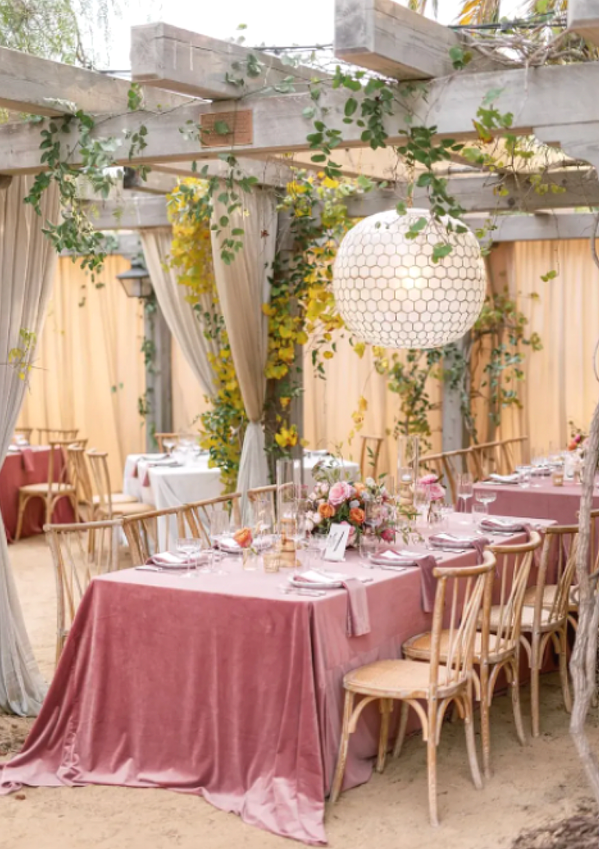 Elegant outdoor wedding reception with pink velvet tablecloths, wooden chairs, floral centerpieces, and hanging lanterns in a decorated garden setting.
