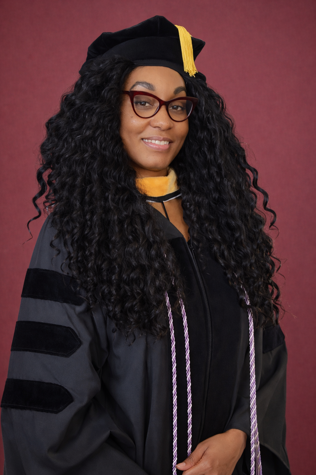 Woman wearing graduation gown and cap, with glasses and long curly hair, smiling against a plain background.