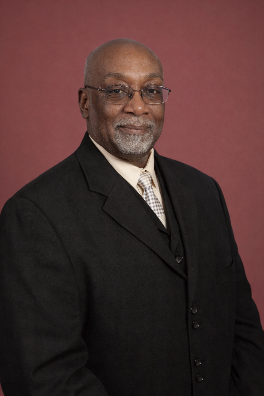 A professional portrait of an older African American man with glasses, salt and pepper beard, dressed in a black suit, white shirt, and patterned tie, standing against a muted pink background.