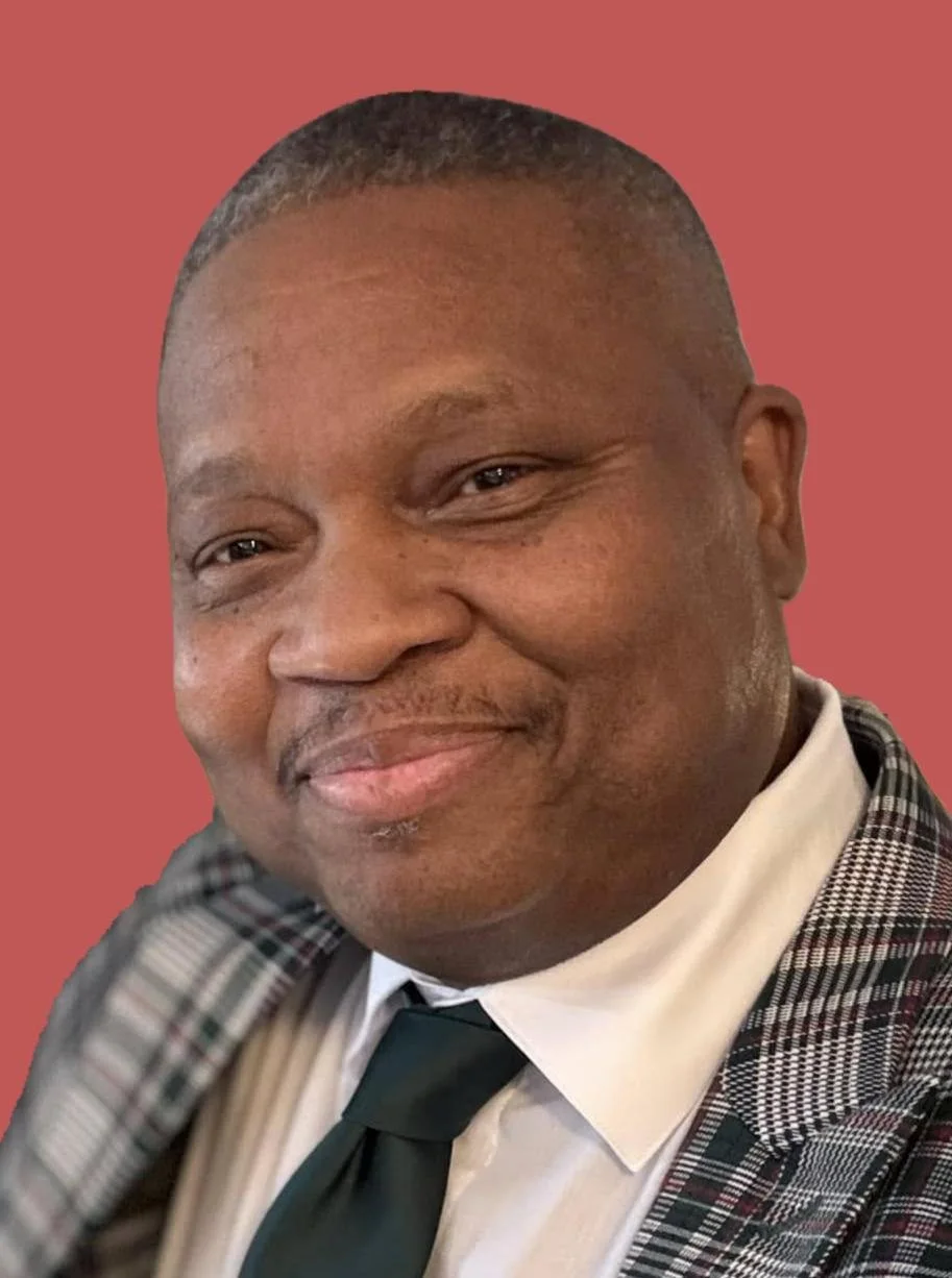 Close-up of a smiling man in a checkered blazer, white shirt, and dark tie, against a solid pink background.