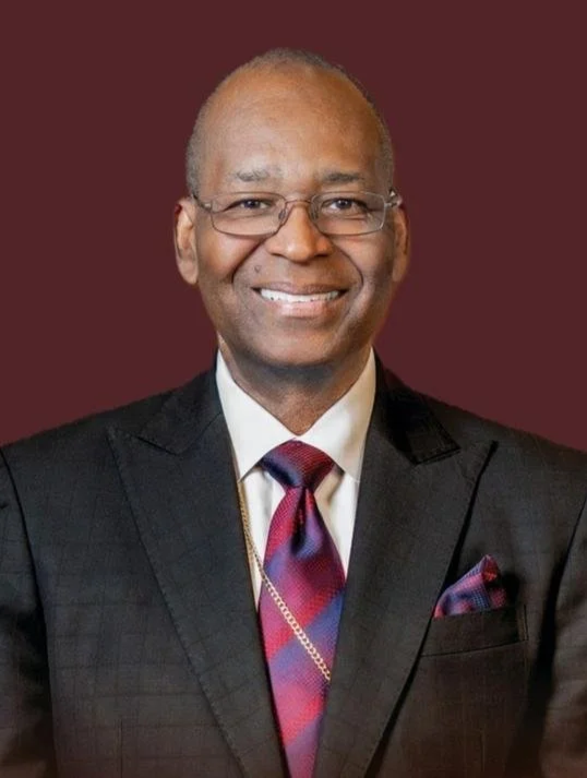 A smiling man in a suit and tie, wearing glasses, with a dark red background.