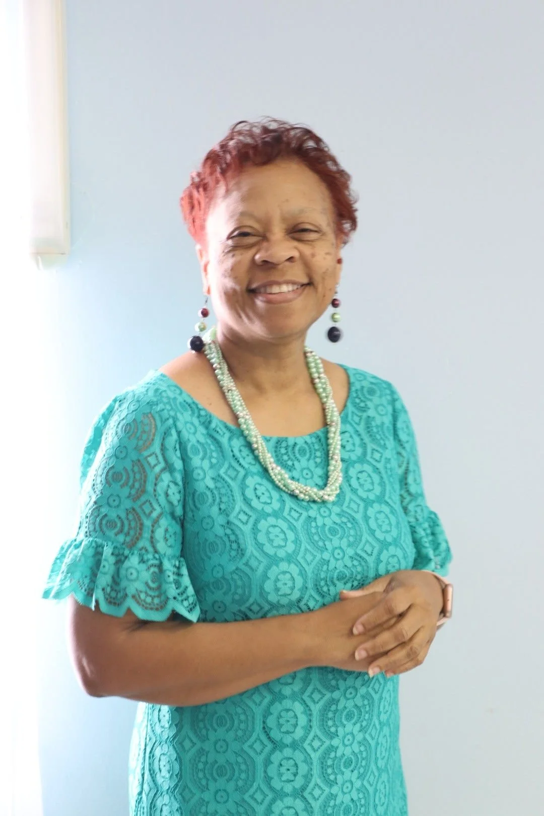 A smiling woman with short red hair, wearing a turquoise lace dress, pearl necklace, colorful earrings, and a smartwatch, standing against a light blue wall.