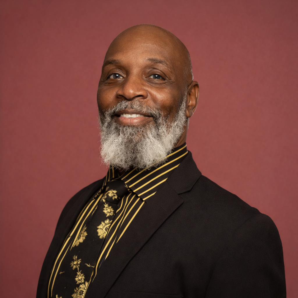 Portrait of a smiling older African American man with a gray beard, wearing a black suit with a striped shirt and a floral tie, against a pink background.