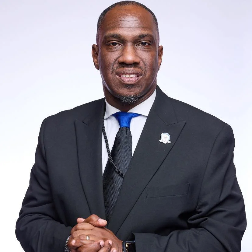 Professional portrait of a man in a black suit, white shirt, and blue tie, standing against a plain white background.