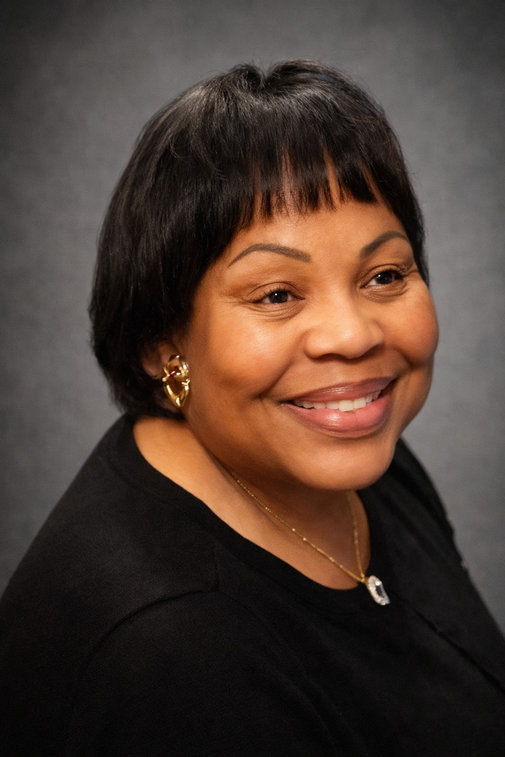 Portrait of a smiling African American woman with short black hair, gold earrings, and a gold necklace against a dark background.
